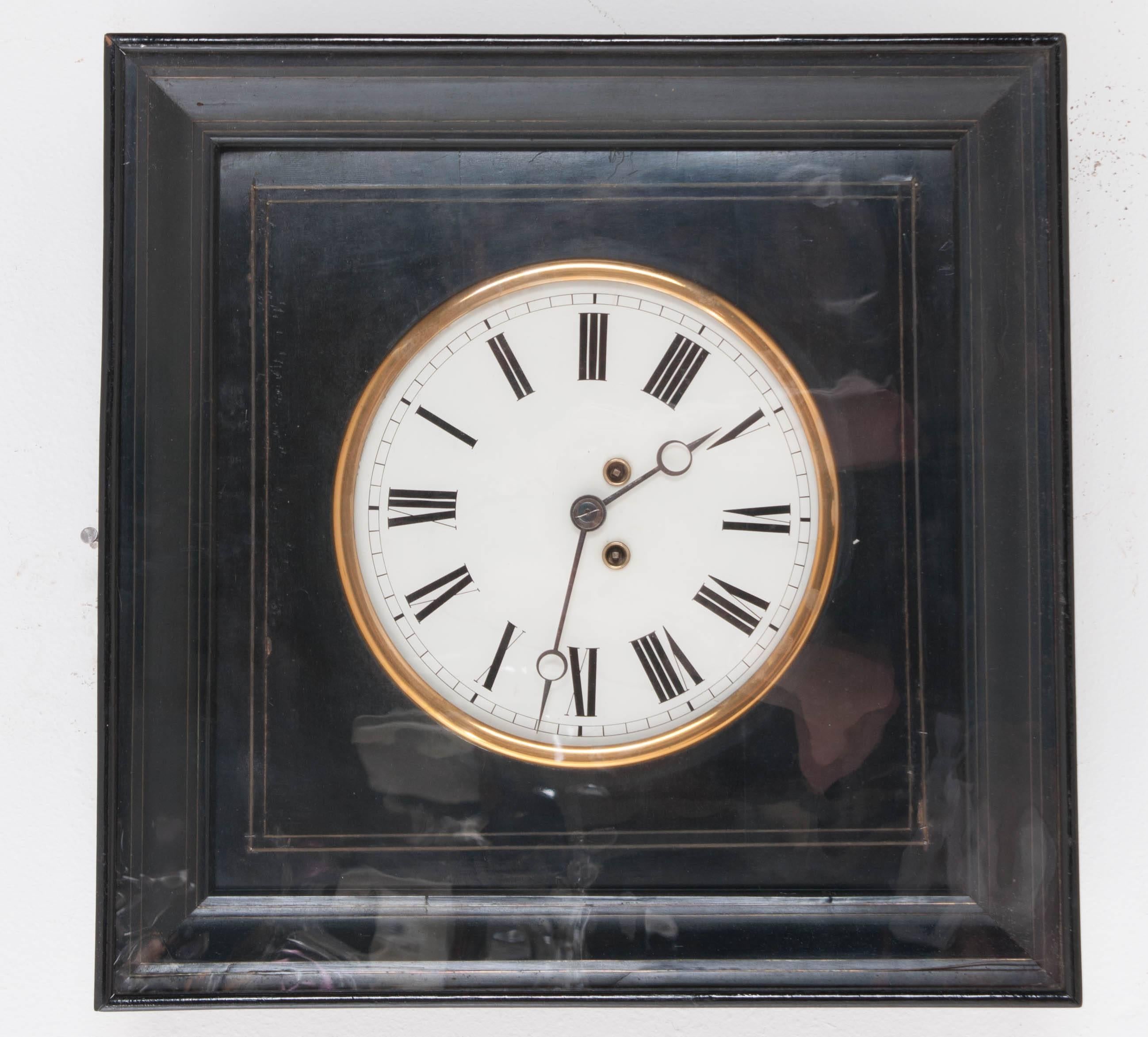 A stunning French wall clock in a square ebony frame, cleaned with its working parts. Surrounded by a brilliant brass ring, the clock's white face stands out vividly against the ebony casing. Thin, inlay brass strips border the interior frame