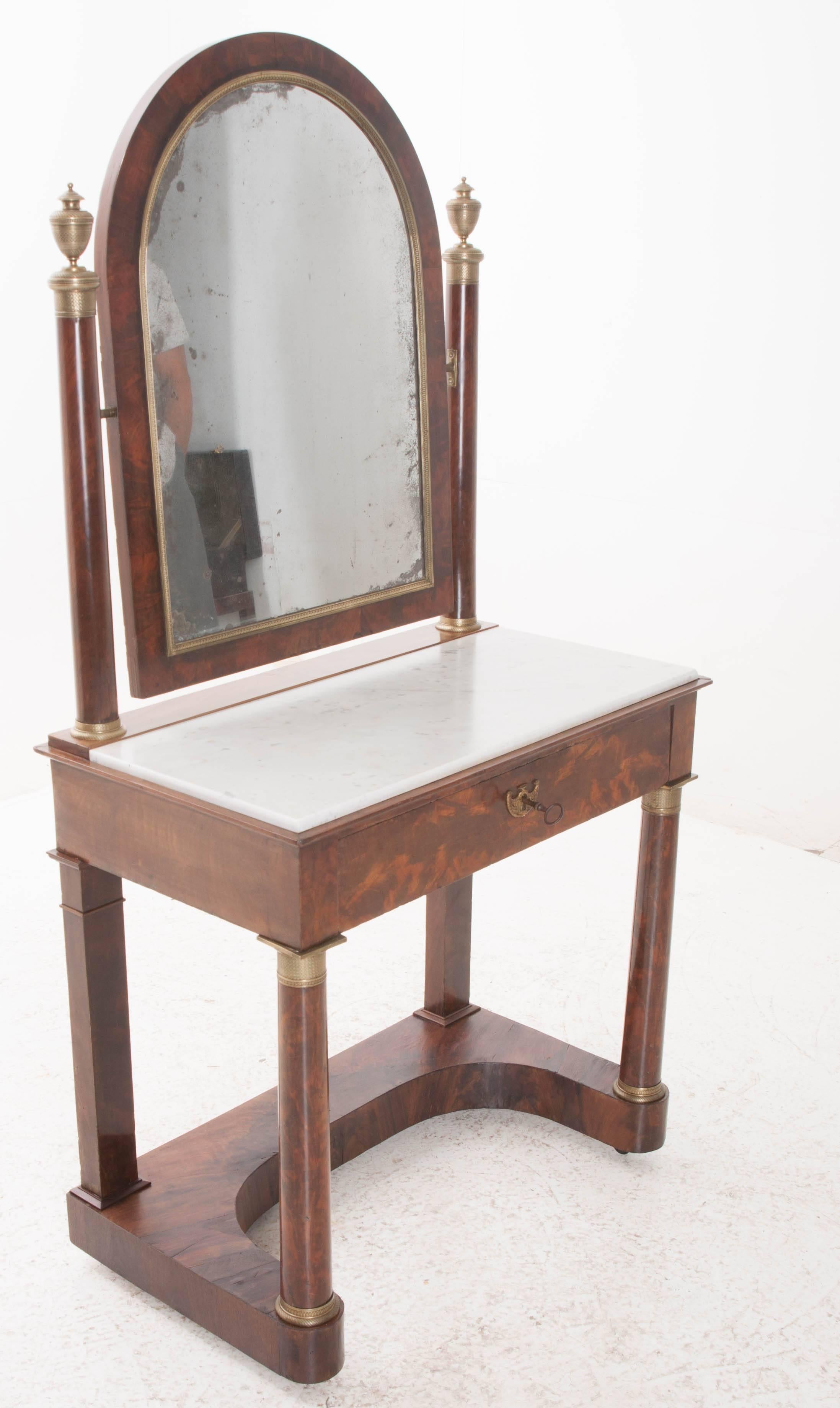 This exceptional French 19th century Empire mahogany dressing table is complete with its original mercury glass mirror and white marble top. The arched mirror boasts brilliant foxing and is framed with beautiful brass trim. It is supported by