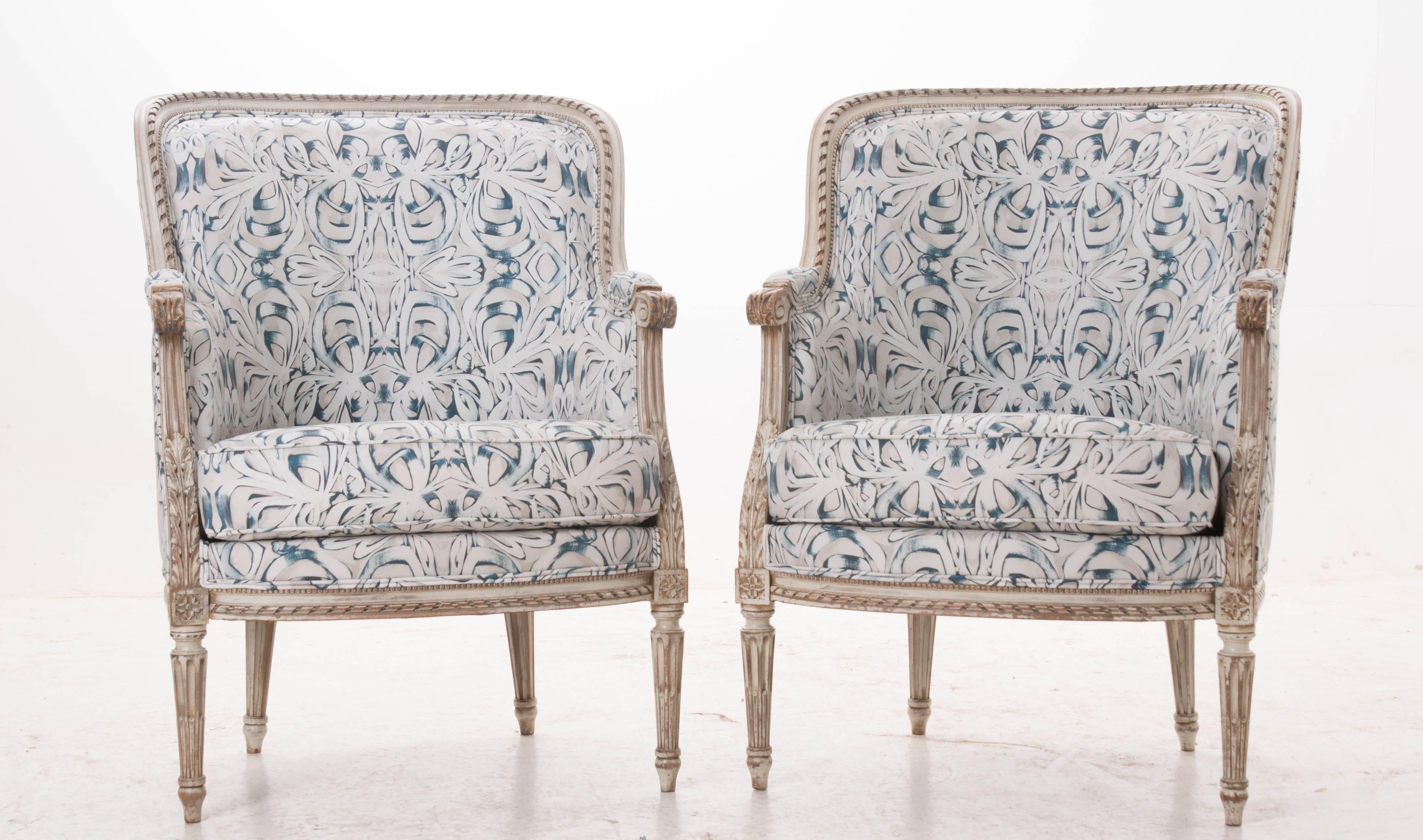 Pair of French barrel back hand-carved and painted bergères upholstered in modern fabric by Amanda Talley of New Orleans, LA. The 'Dungarees' pattern by Talley is the perfect paring for the worn cream paint on the antique chairs. The chairs are