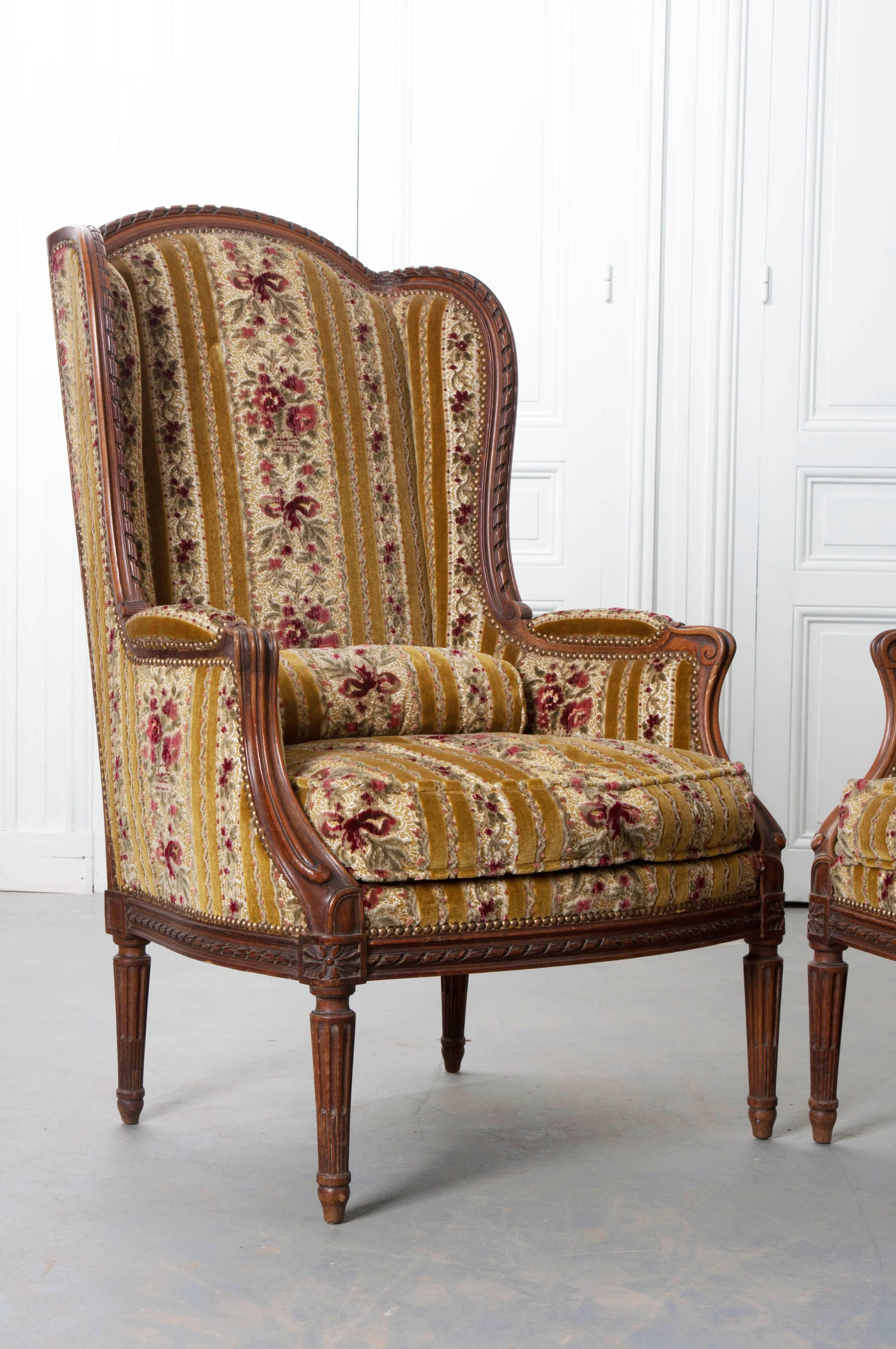 A pair of brilliantly carved French walnut bergères that combine elements of Louis XV and XVI styles. The back features a twisted ribbon motif that can also be found on the chairs' rails. The arms have a more fluid, scrolled design, influenced by