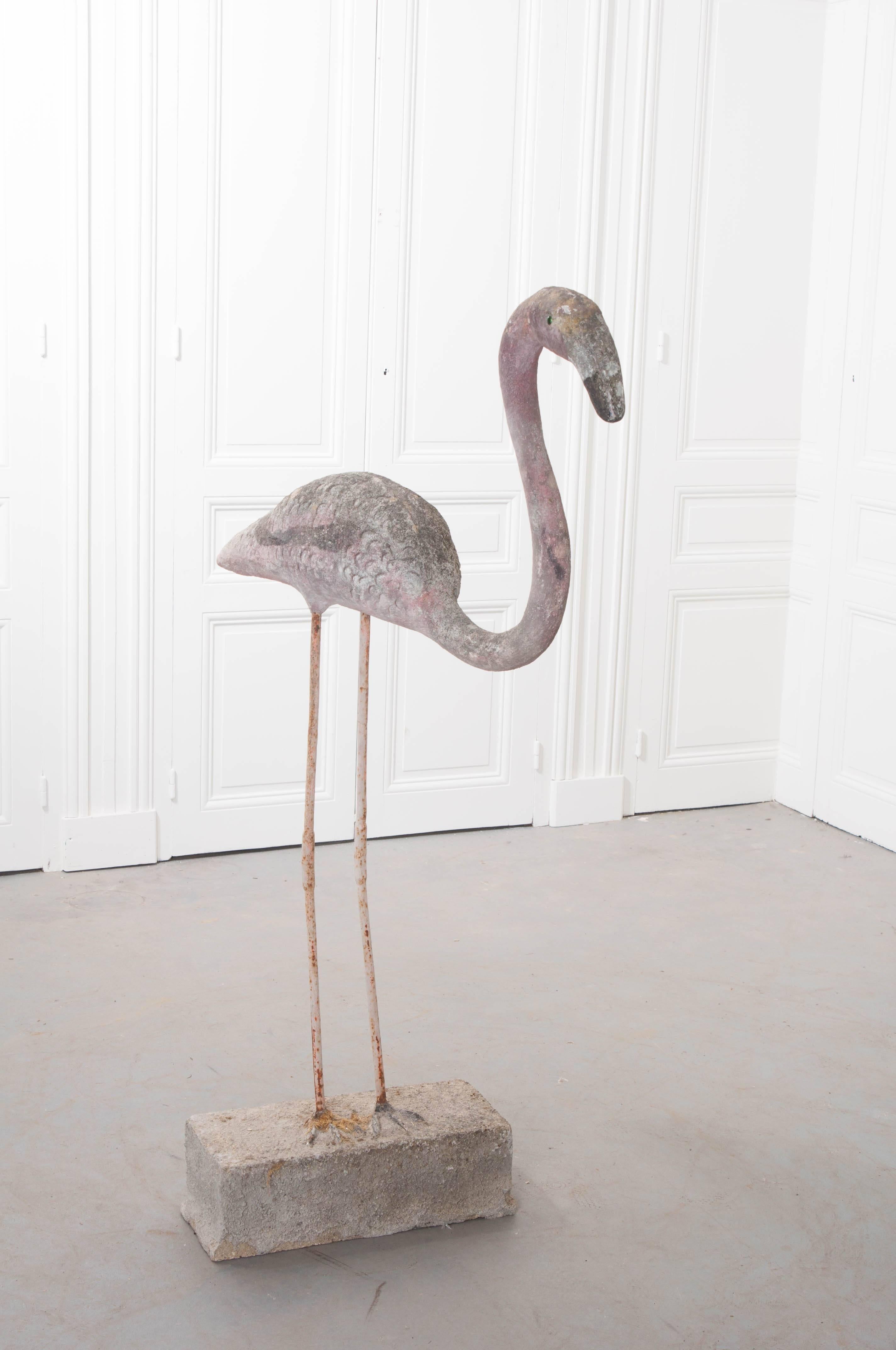 A majestic reconstituted stone flamingo perched upon a stone block with legs made of iron from England, circa 1920. Its graceful, curved neck and detailed feathers create a striking resemblance to the beautiful blush birds. The paint has weathered