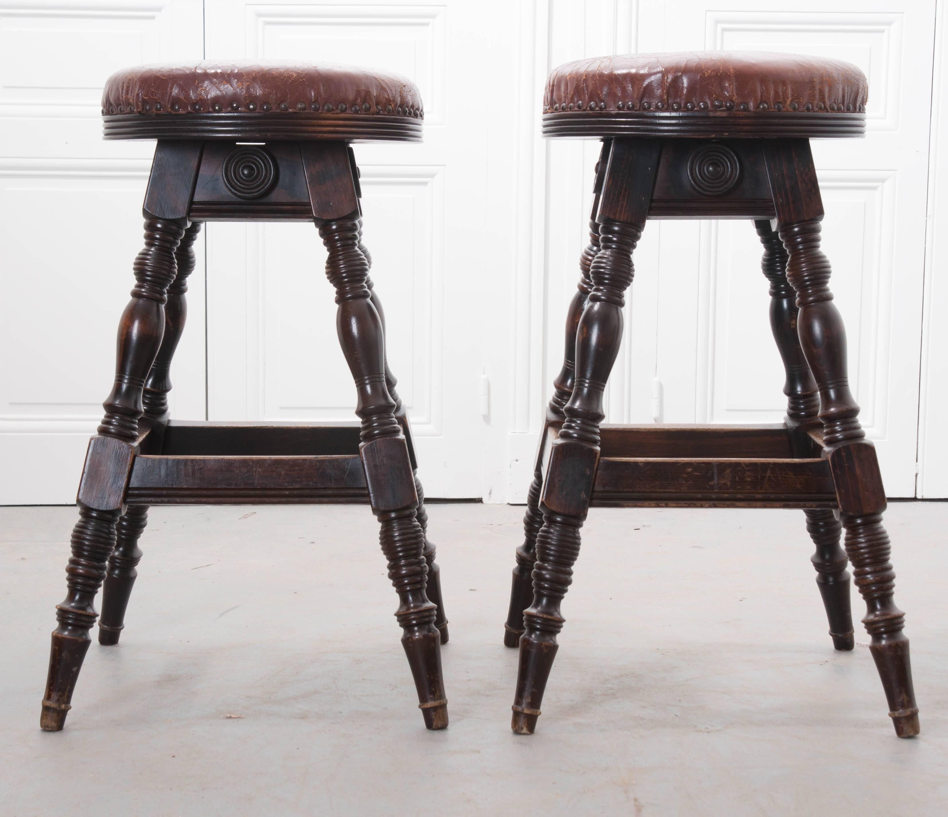 A handsome pair of antique oak pub stools with leather seats from the end of the 19th century, England. These stools have worn, cordovan leather seats that are secured to their frames using bronzed nailheads. The seats are fixed and do not swivel.