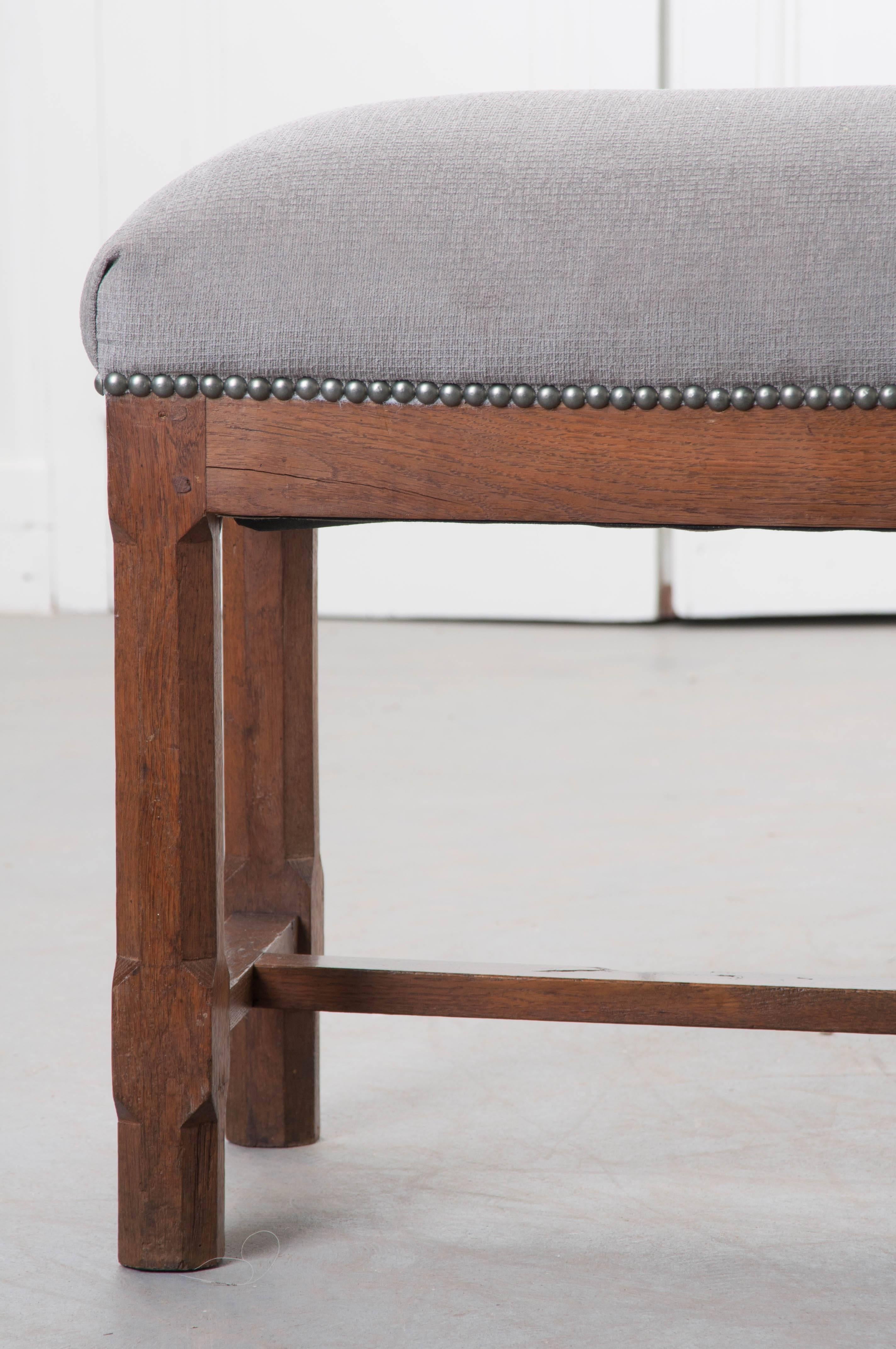 A newly upholstered antique French bench from the 19th century. The Provincial bench is made of solid oak that has been fixed together using peg and hole joinery. The chamfered legs are joined by an I-stretcher below. The recent grey upholstery is