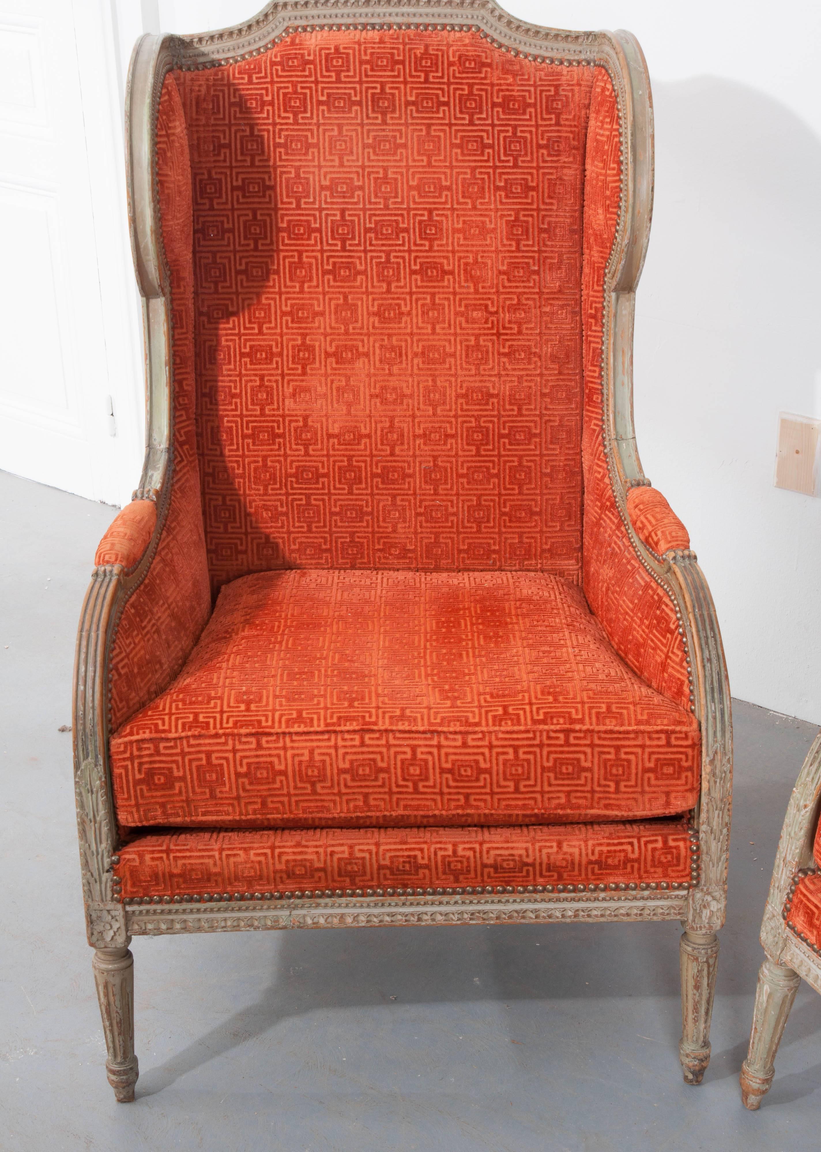 A fine pair of French Louis XVI bergères from the 19th century. The chairs are upholstered in a vivid flame persimmon colored cut velvet, with a geometric pattern, and affixed to the frame with nailheads. A remarkable aged patina has consumed the