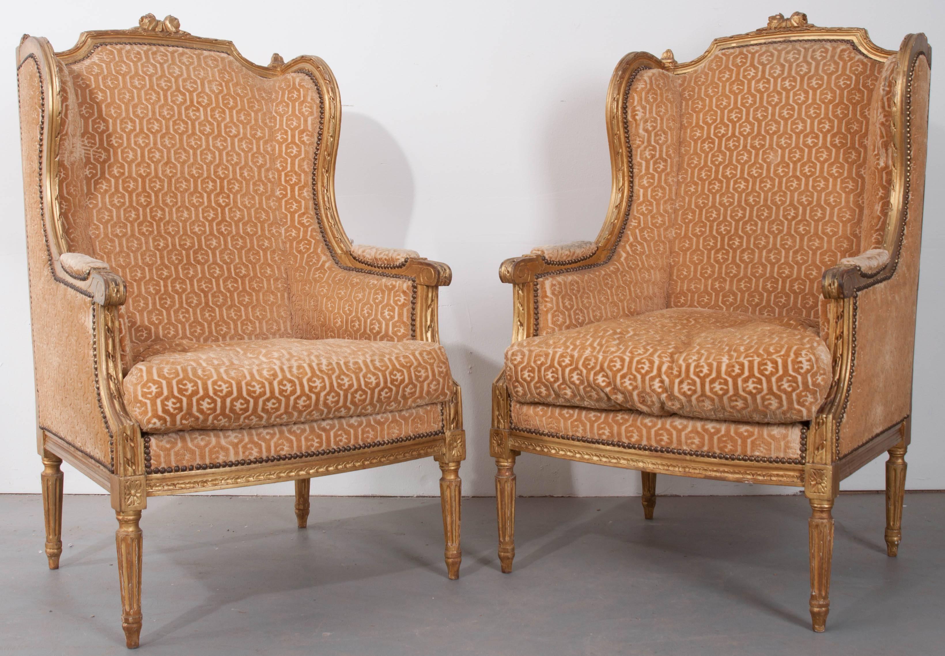 A marvelous pair of gold gilt Louis XVI wingback bergères from 19th century France. These beautiful chairs are upholstered in a gold cut velvet, done in a geometric pattern, and secured to the frame with brass nailheads. The gilt frame is lustrous