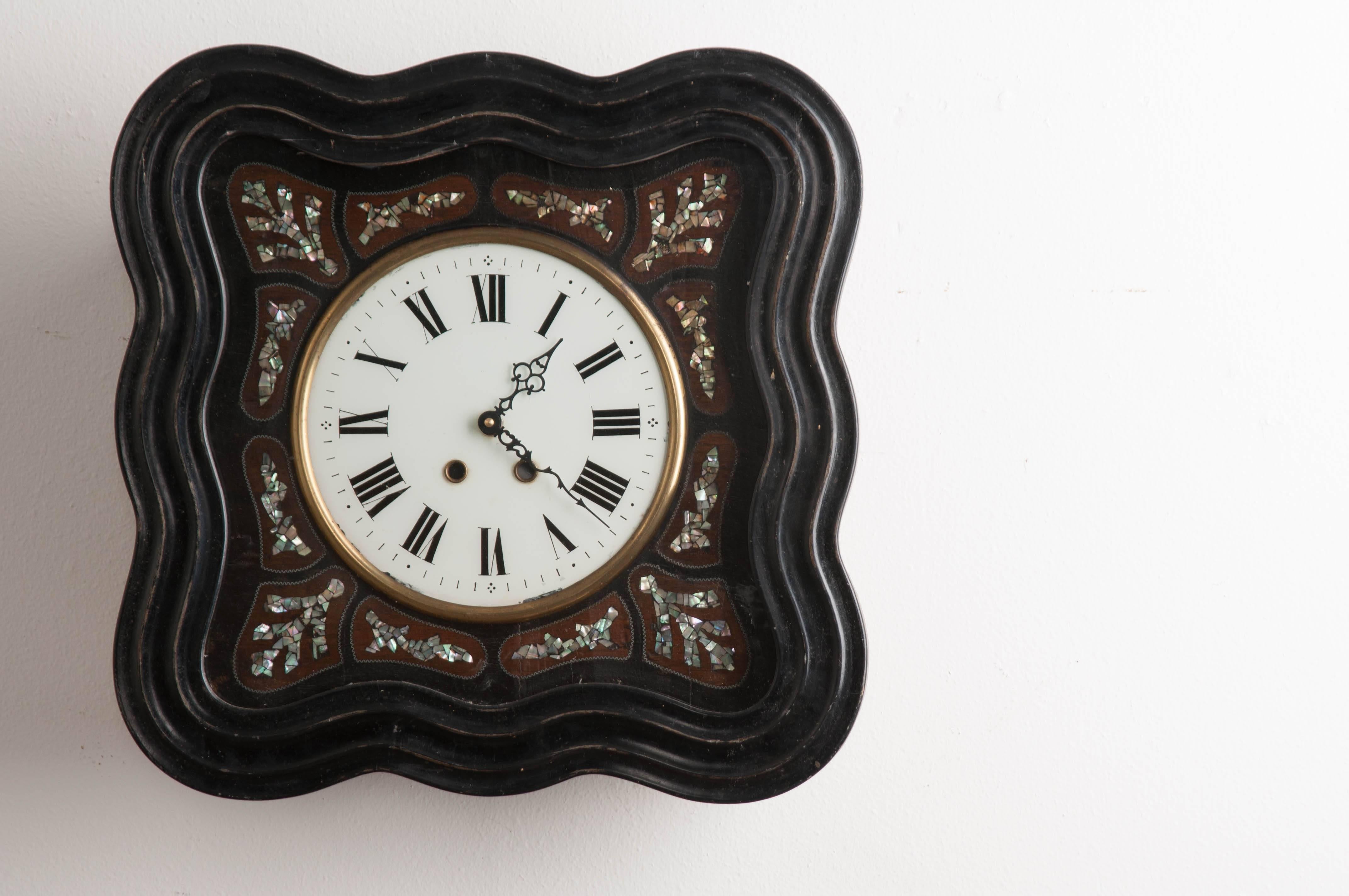 A Napoleon III style wall clock, with exceptional mother-of-pearl inlay, from 1850s France. This shapely clock is square in shape, with a wonderfully wavy frame. This frame is hand-carved and ebonized. It closes before a hand-painted clock face,
