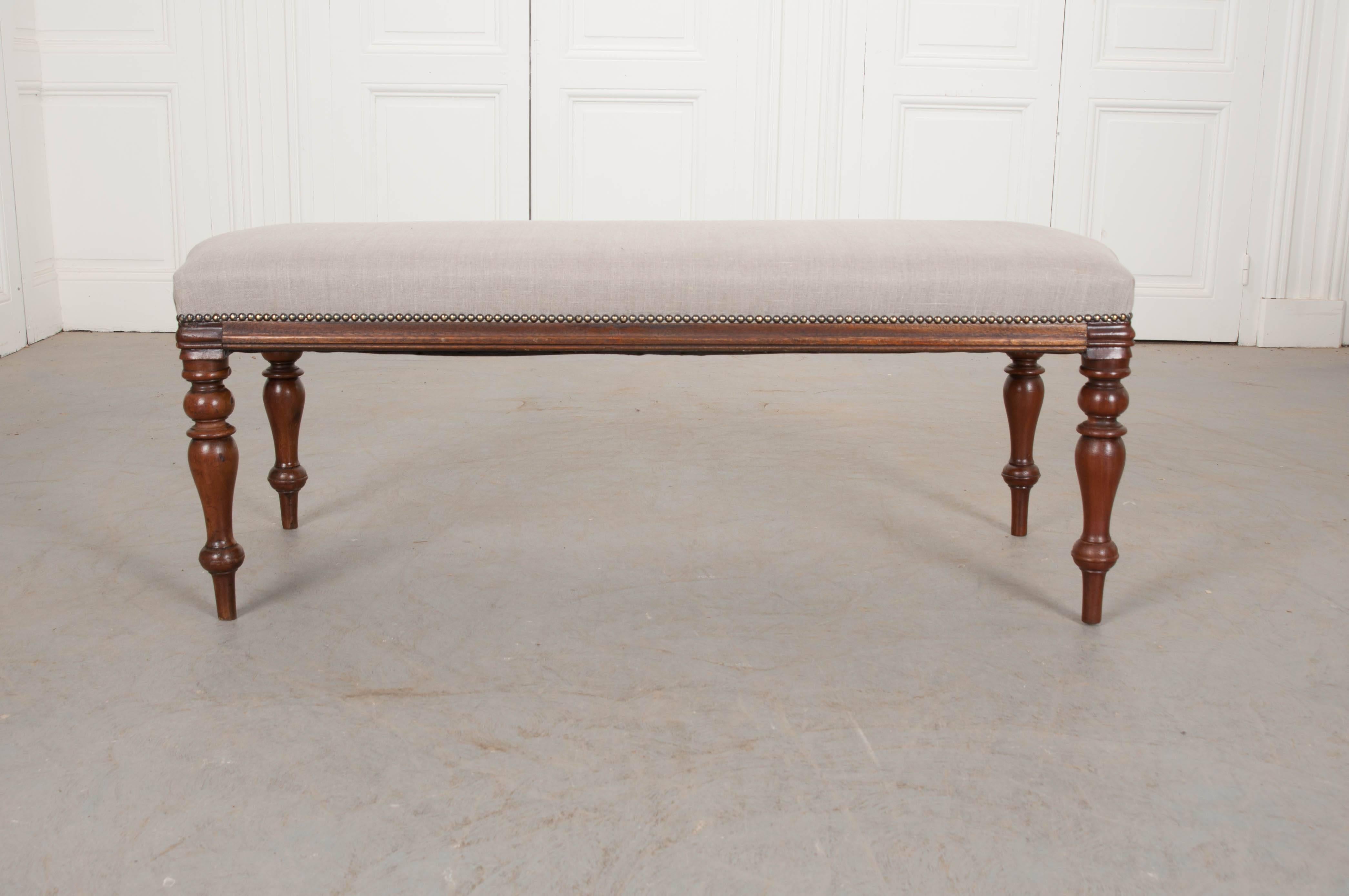 A newly upholstered bench from the end of the 19th century, England. This long, mahogany bench is upholstered in a taupe-colored woven linen with natural fibers that is affixed to the frame using antiqued metal nail heads. The bench is lifted on