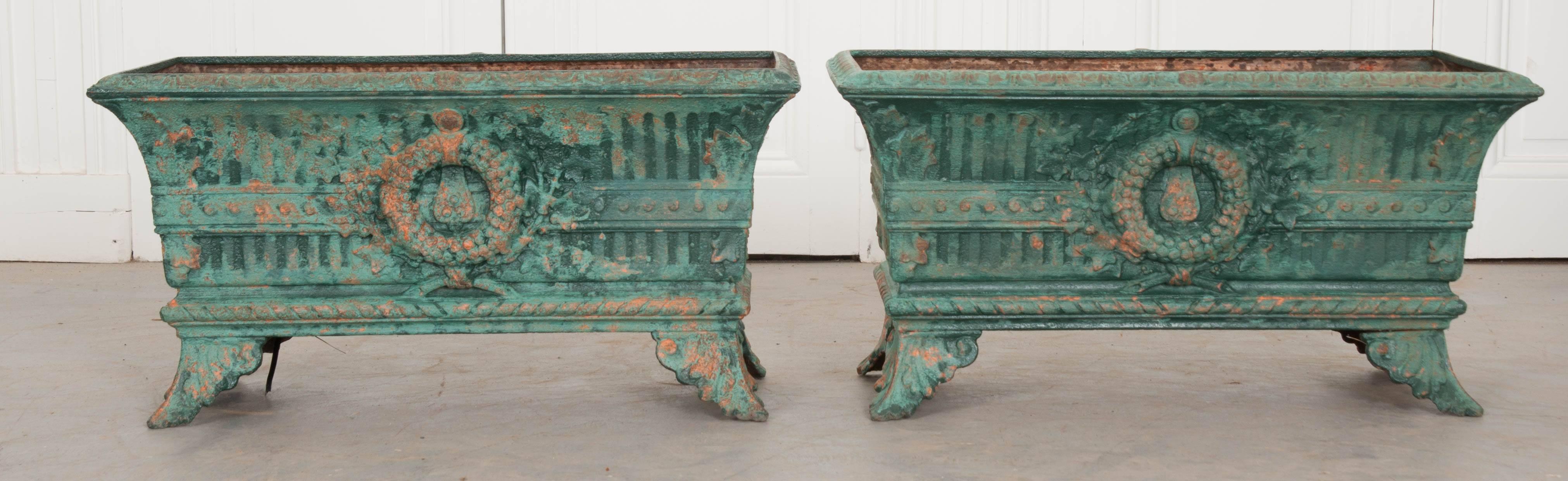 Two incredible French cast iron planters that have been painted, circa 1870. The rectilinear planters are perched atop decorative bracket feet, don in an acanthus leaf motif. Front and center lie circular wreaths that lie on top of a belt encircling