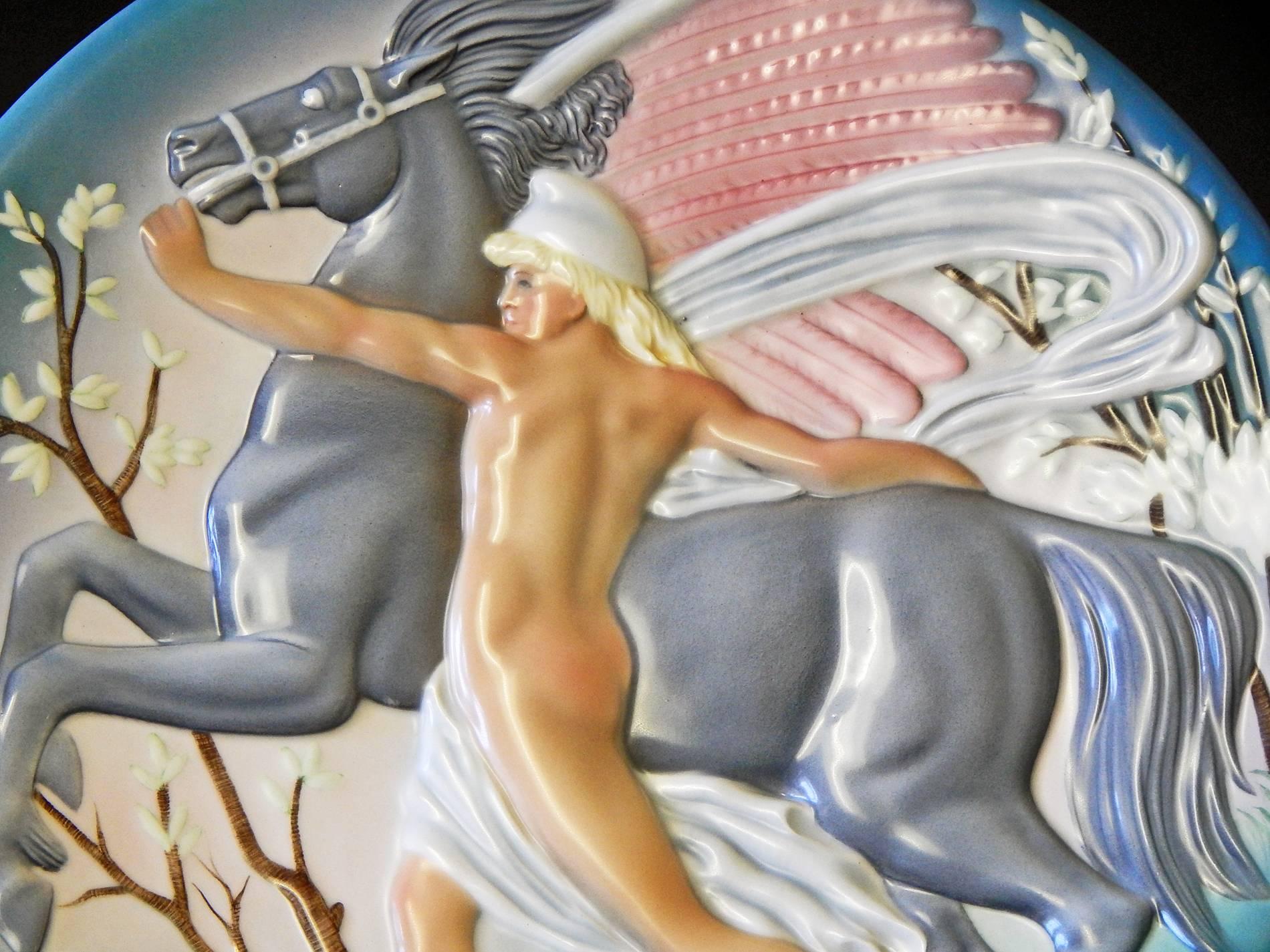 Magnifiquement et subtilement émaillée dans des tons de gris, vert, rose et beige, cette étonnante rondelle sculpturale Art Déco représente Bellérophon, une figure masculine nue de la mythologie grecque, retenant un Pégase ailé dans un paysage