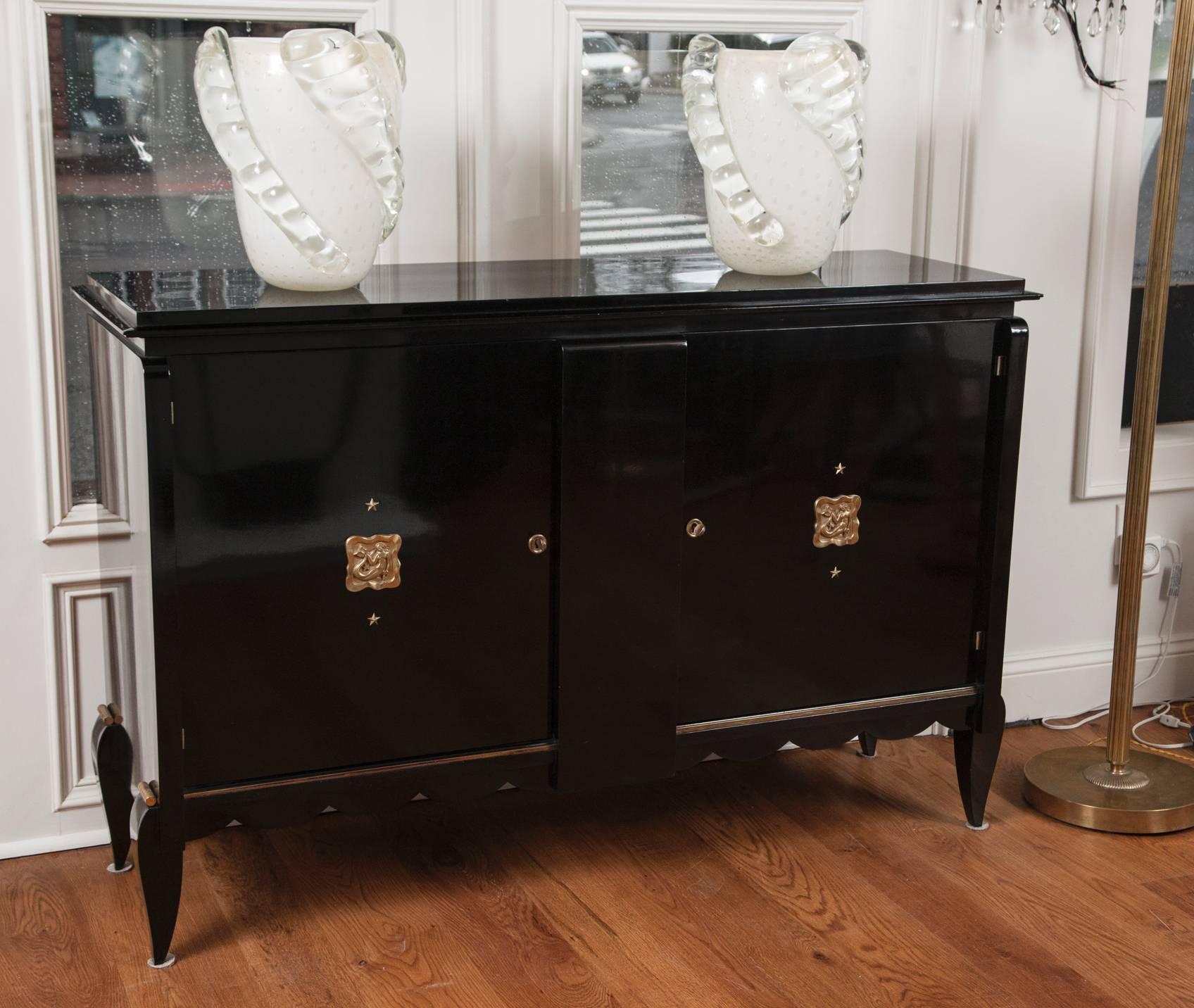 Gorgeous ebonized French sideboard comprised of two doors with curved legs and a scalloped apron, adorned with lovely brass mermaid and star placards, in the style of Leleu, circa 1940. Lacquer hand rubbed.  Interior outfitted with shelves in a
