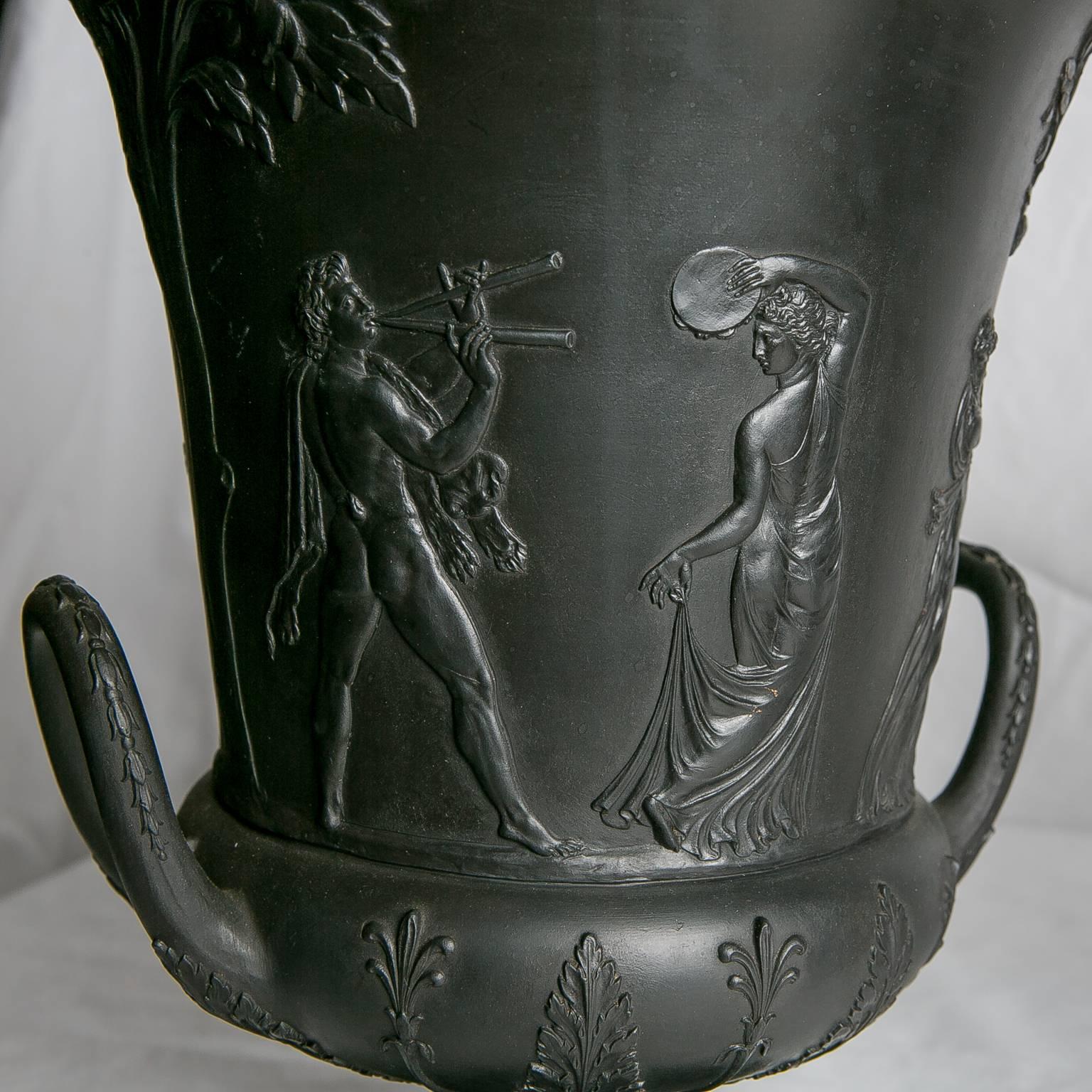 A pair of mid 19th century Wedgwood Black Basalt campana shape urns with neoclassical decoration of figures and a design of anthemion and palmette leaves.The urns have simple upturned loop handles. The figures excellently carved in bas-relief