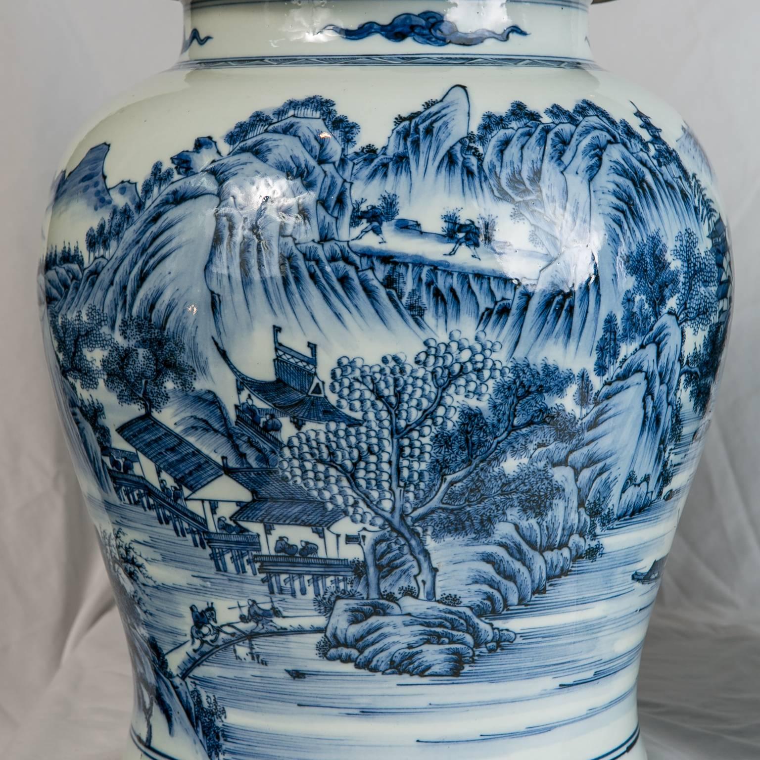 A massive pair of antique Chinese Blue and White ginger jars decorated with an all around mountainside scene. The scene is set in springtime with travelers walking across a bridge ascending up the mountains. Flowering trees, boats on a river, and
