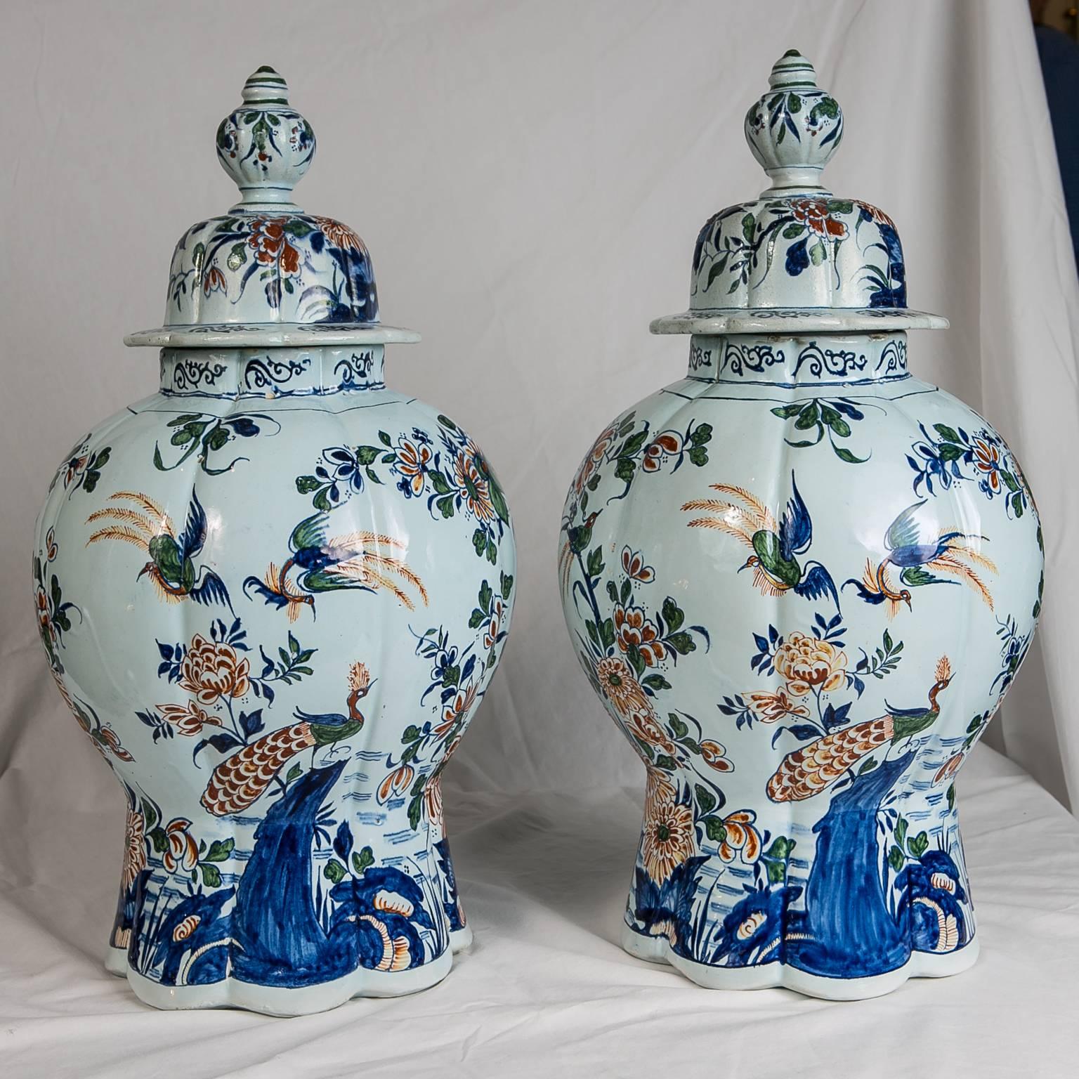 A pair of Dutch Delft ginger jars painted in a polychrome palette of strong orange, green, and blue with touches of yellow. The jars are decorated with a beautiful all around scene showing long-tailed birds with blue wings, a green breast, a red