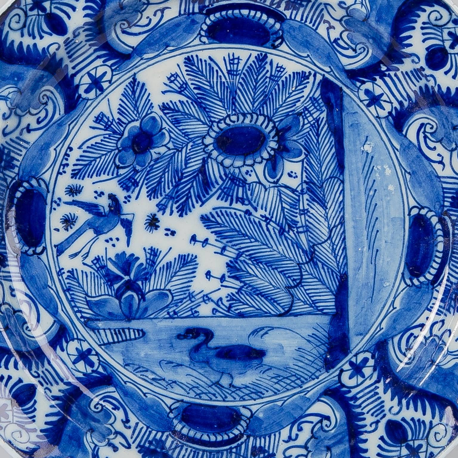 A Dutch Delft 10 inch charger where the center shows a beautiful, lively, and very detailed ducks-in-a-pond scene with giant flowers overhanging the edge of the pond, a duck swimming, and a long-tailed bird flying above. The scene and the