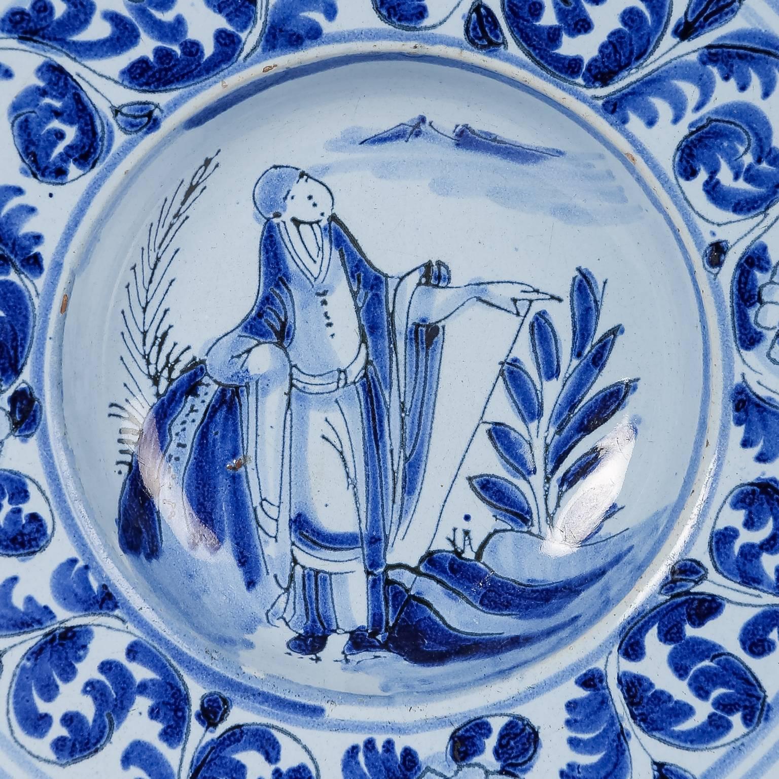 We are pleased to offer this small blue and white Dutch Delft plate decorated in shades of cobalt blue, showing a traveler dressed in Chinese monastic robes with a walking stick, made circa 1770.
The wide border is decorated with scrolling leaves