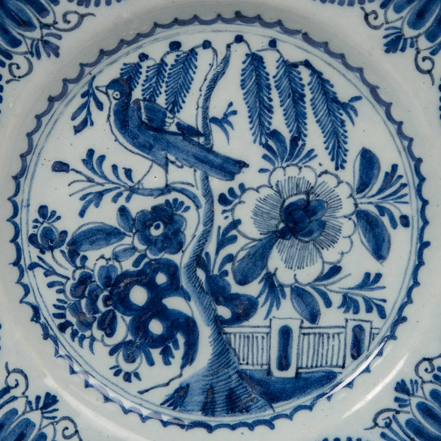 An intricate chinoiserie blue and white Dutch Delft dish painted in cobalt blue showing a garden scene with a long tailed bird perched on a branch of a plum tree, an oversized peony, rockwork, and a garden fence. The lovely design of the bird in the