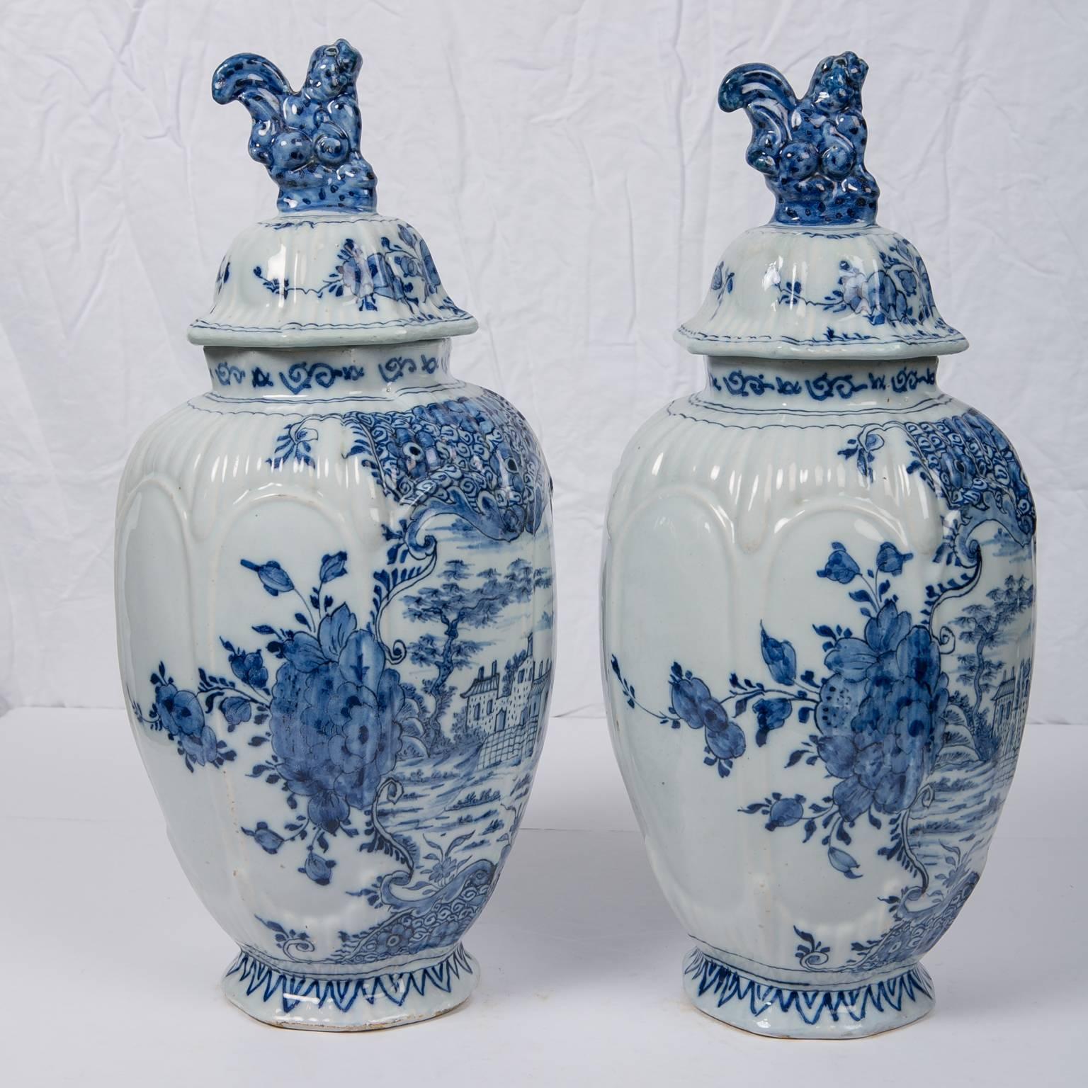 A lovely pair of antique Blue and White Delft jars. The covered jars are ribbed with panels painted in a soft cobalt blue showing a Dutch village. At the sides of each scene are lush oversized peonies.
Each vase is topped by a traditional cobalt