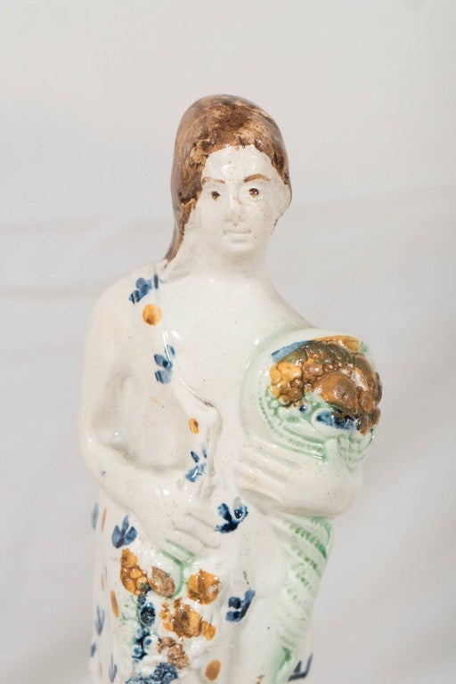 We are pleased to offer this naive and charming 18th century creamware figure of a woman personifying summer. Modeled standing on a plinth she holds a cluster of grapes, and a cornucopia filled with fruits and vegetables. The attractive base is