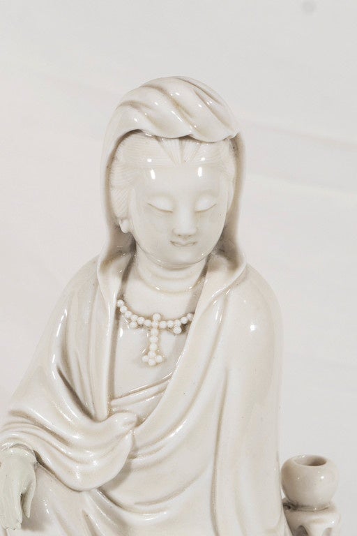 A fine Dehua Blanc de Chine figure of Guanyin under a rich ivory toned glaze. The goddess is clothed in a voluminous flowing robe.which is open at the chest to reveal a beaded necklace. Her face is lowered with a benign expression. The Guanyin is a