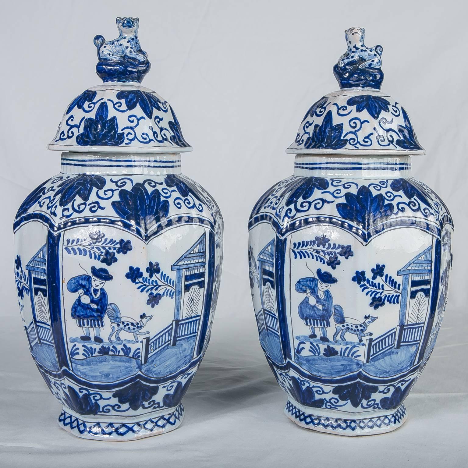 A pair of Dutch delft covered jars with panels painted in deep cobalt blue showing a chinoiserie scene of a boy and his dog walking near a pagoda.
The covered jars topped by traditional leopard finials.
Made in the Netherlands in the first half of