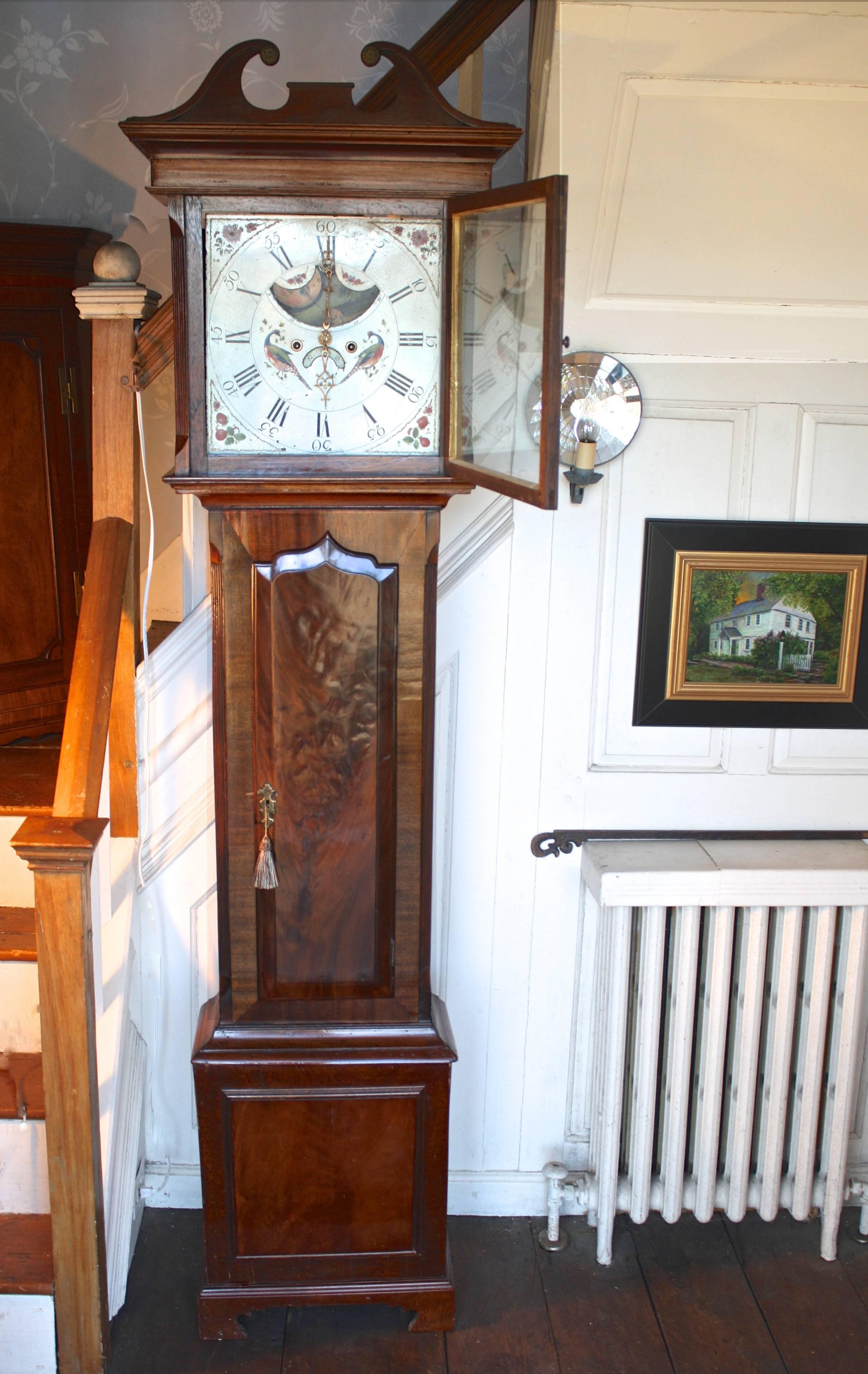 A seven foot tall Regency period clock with a finely detailed mahogany case and paint decorated face. The mahogany case includes solids as well as crotch and flame veneers, with wide grained bandings.  The complex elements of its face paint