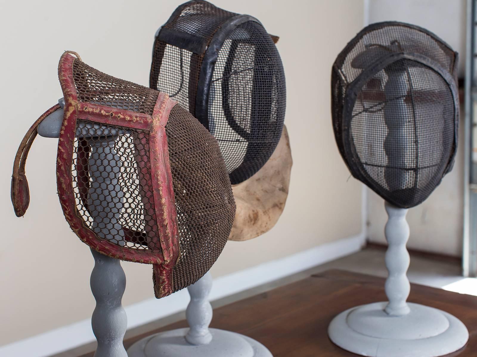 The unique shape of these antique French fencing masks, circa 1910 with their wire mesh protective guards set upon heavy iron frames are here displayed effectively upon painted wooden wig stands from 19th century, France. Each French fencing mask