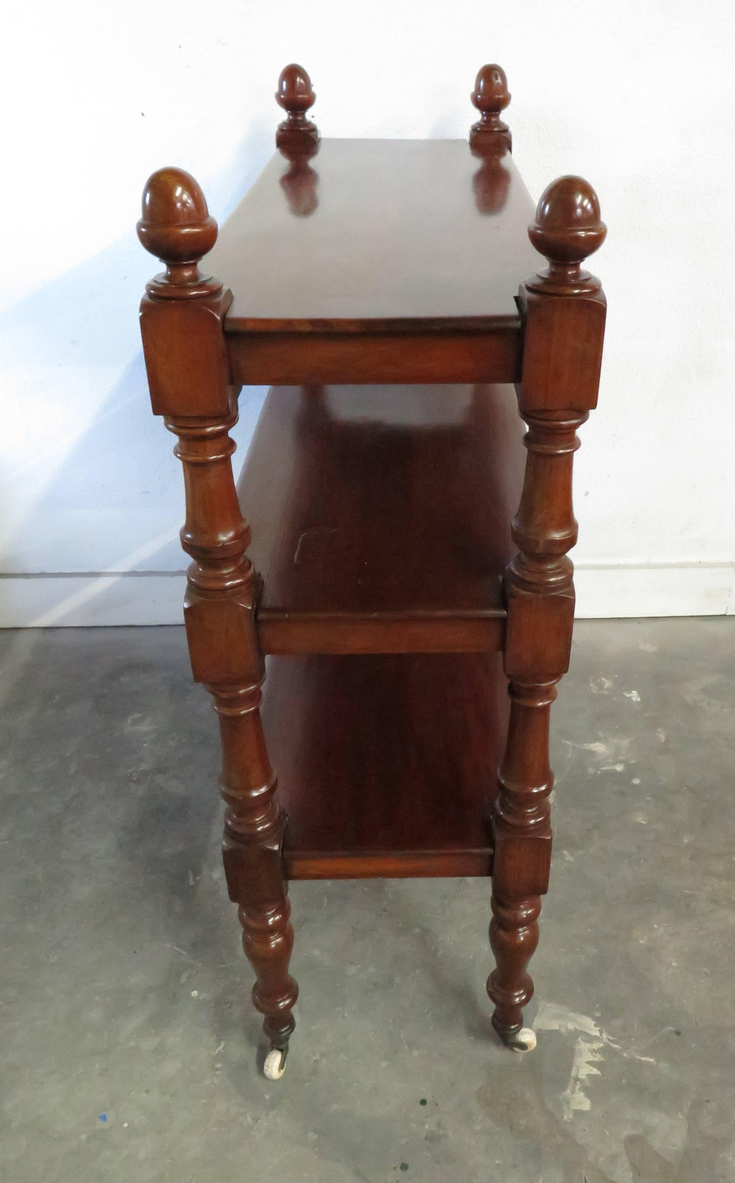 Antique English mahogany cart featuring three tiers and decorative turned posts, set upon rolling casters.