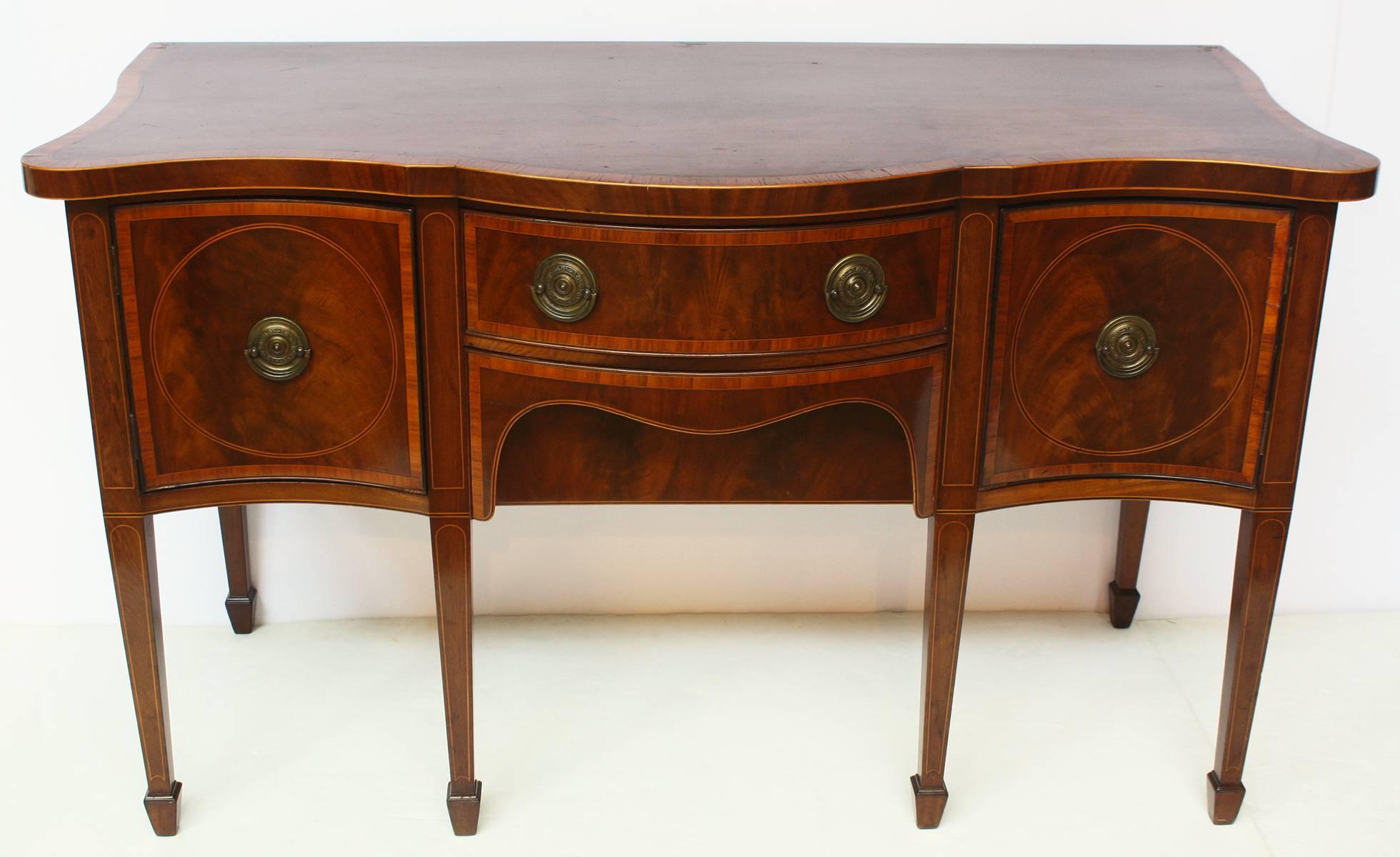 An English George III period mahogany sideboard inlaid in ebony and satinwood, with a bow fronted central drawer flanked by two serpentine fronted hinged doors each containing fitted pull-out trays. All rests on six square tapered legs and spade