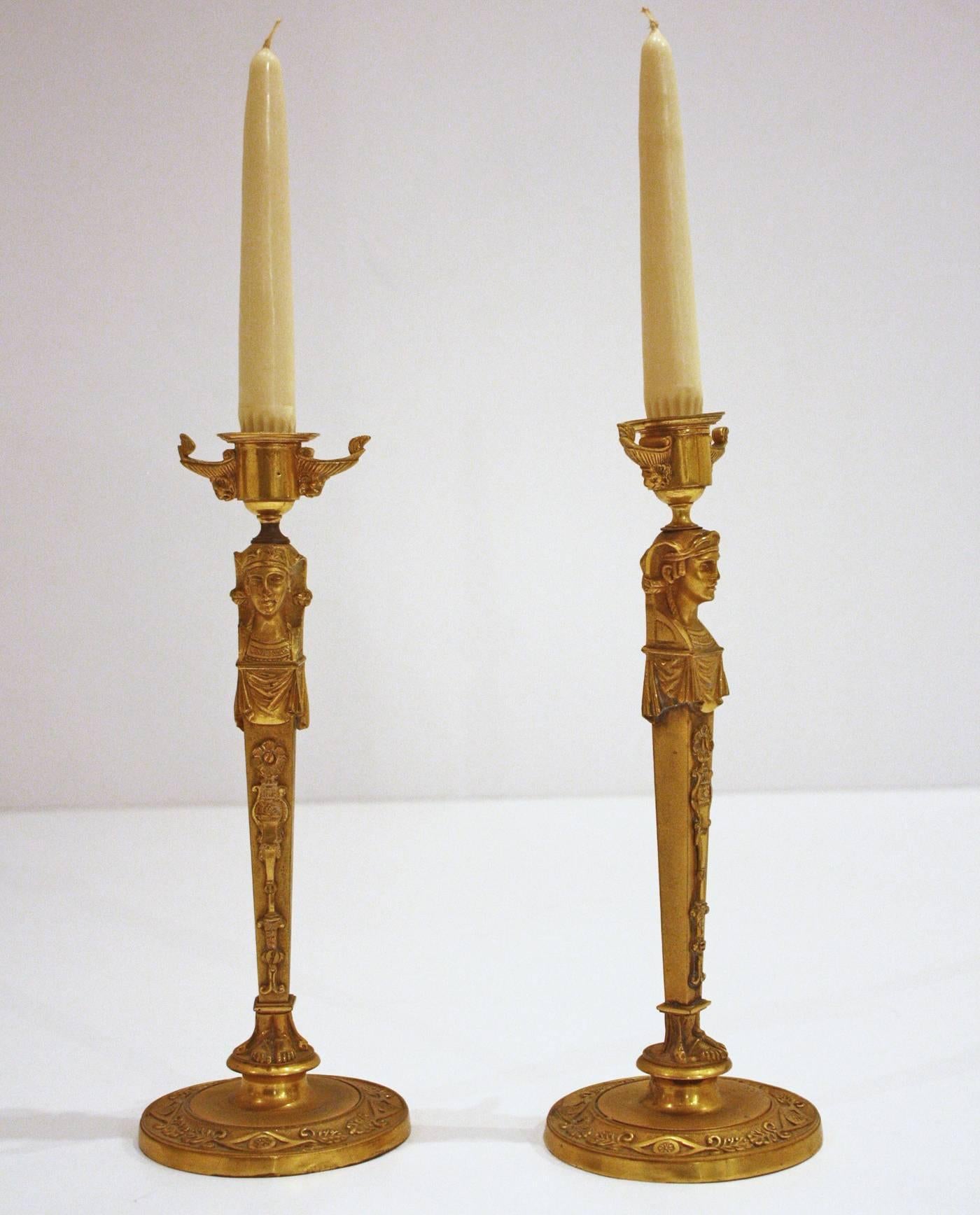 A pair of gilt bronze candlesticks as standing caryatid figures with separate drip pans. Below the drip pans are a pair of handles with lion mask supports. The vase shaped candlesticks have an applied detailing on the front. Below the vase shape are