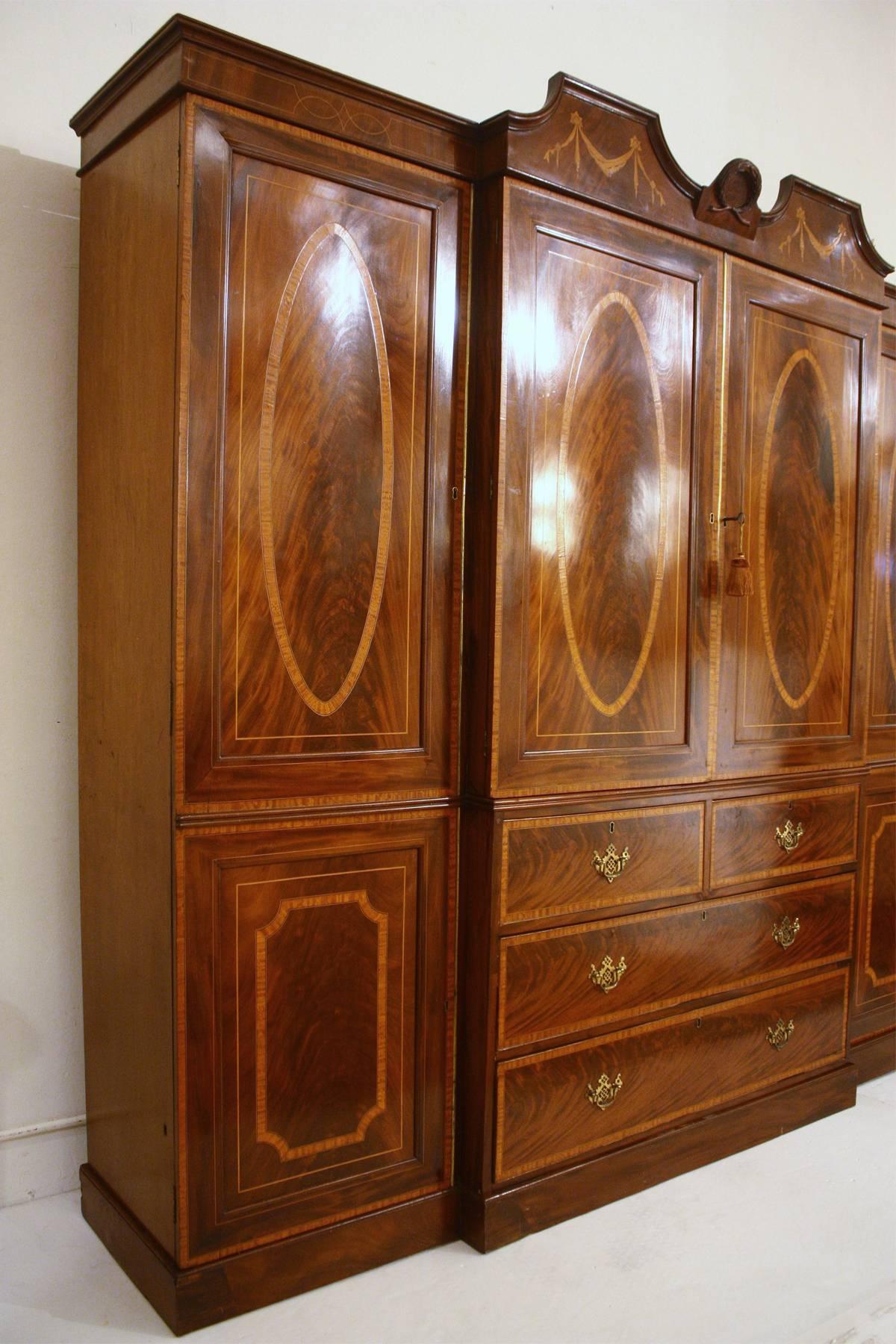 A stunning quality Georgian Revival breakfront mahogany gentleman’s wardrobe/press. The cornice having a broken pediment with a craved laurel wreath in center and two inlaid garland motifs. The central twin doors opening to reveal five oak dressing