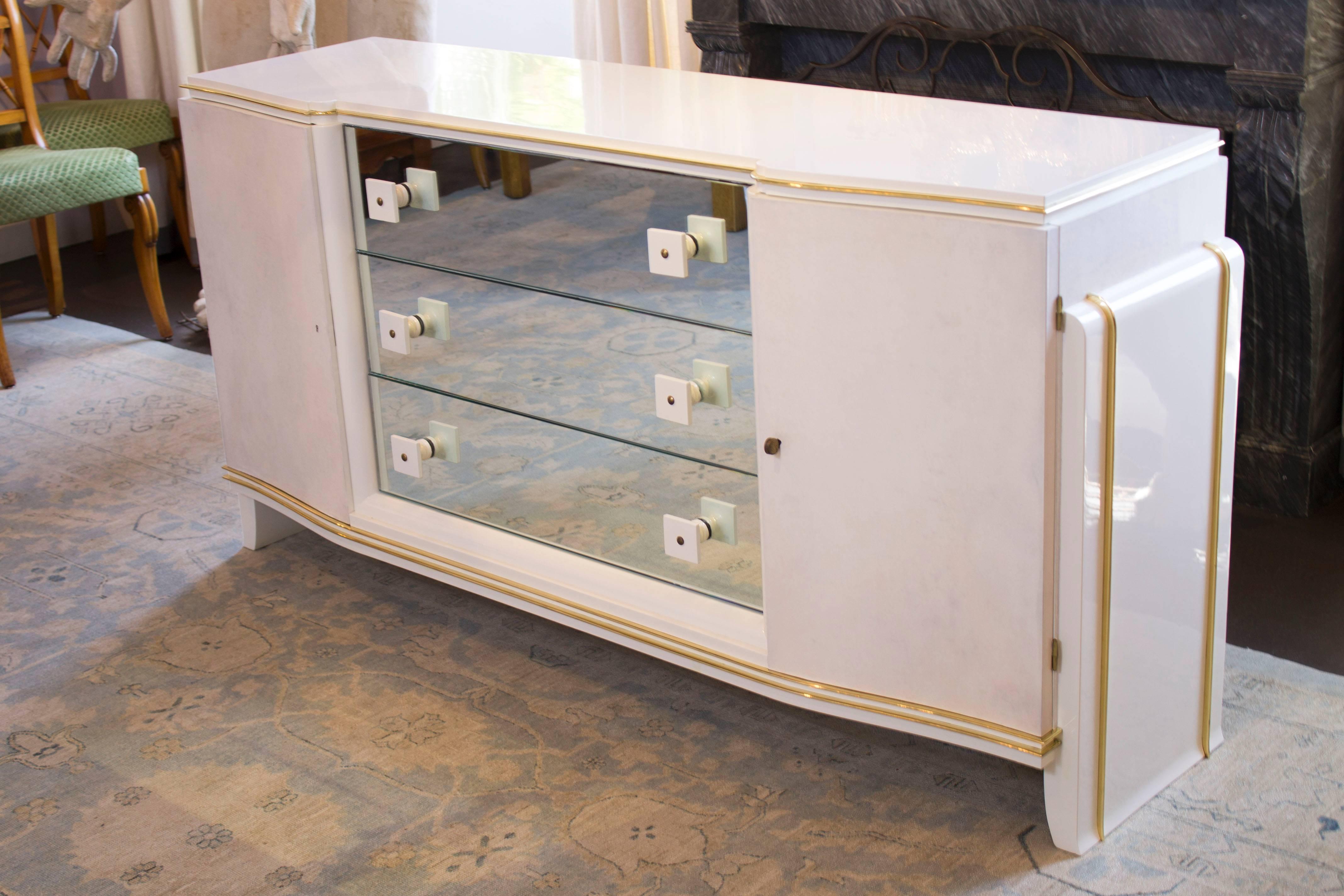 A beautiful white lacquer dresser with brass inlays. It features mirrored drawers and cream parchment leather lined cabinet doors.