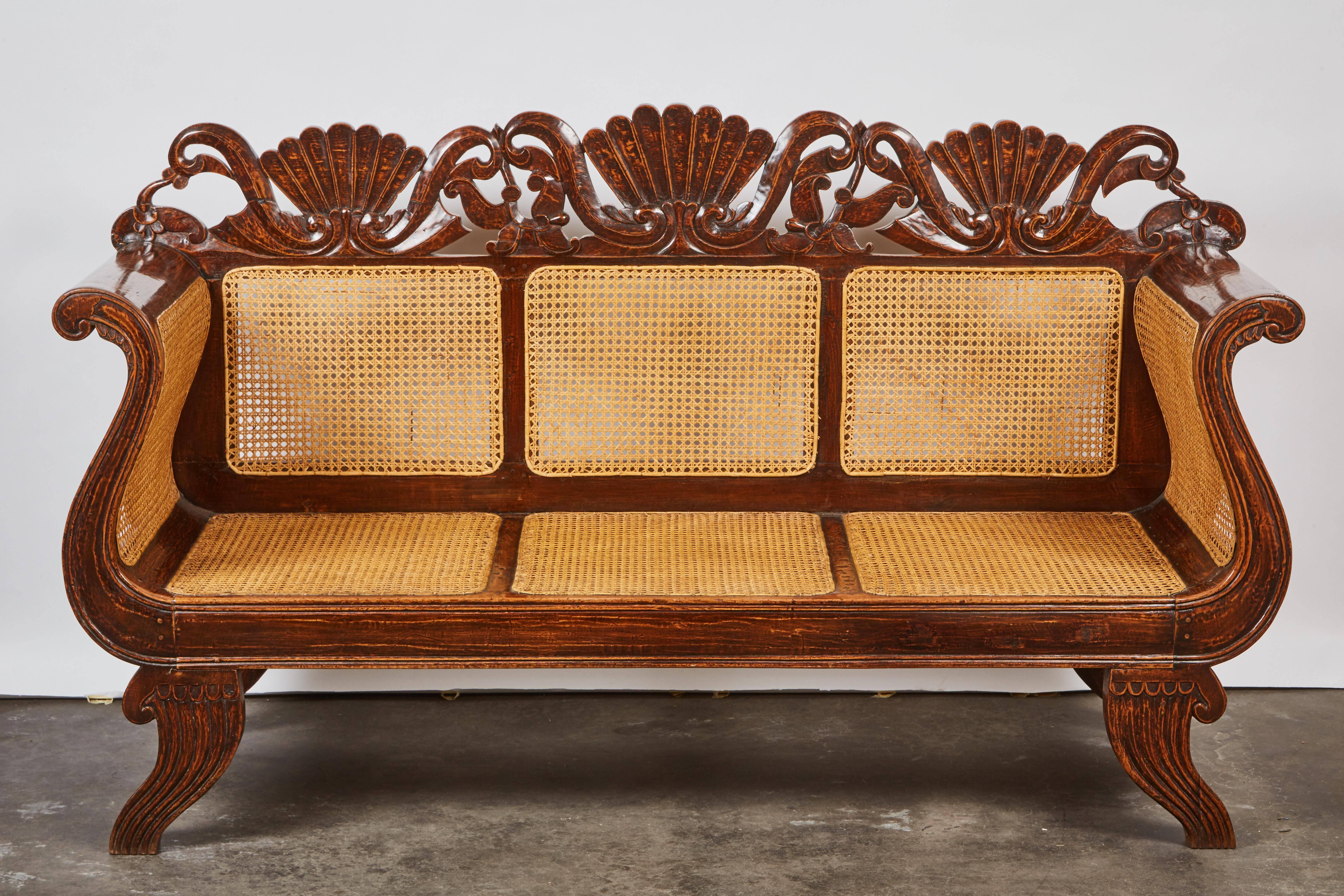 A beautiful Indonesian teak settee with a crest rail featuring carved rocaille or foliage motifs, large in scale. The back features three inset caned panels above a wicker caned seat. Dutch colonial.