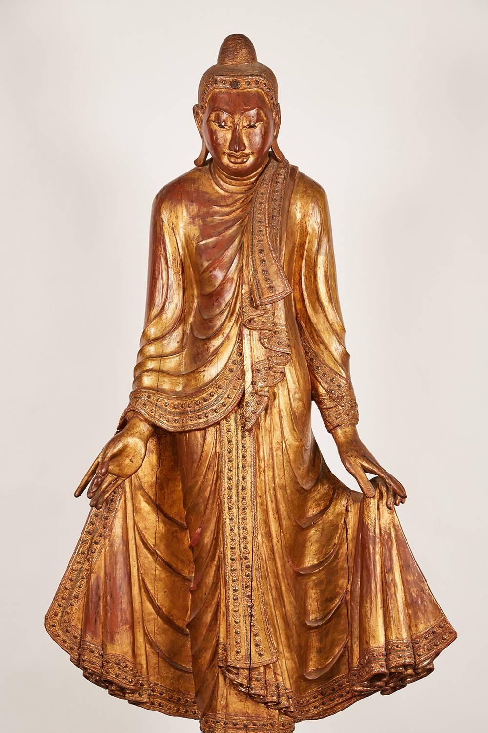 A very large gold Thai standing Buddha featuring intricate inset glass detailing. Supported by a later custom wooden stand
