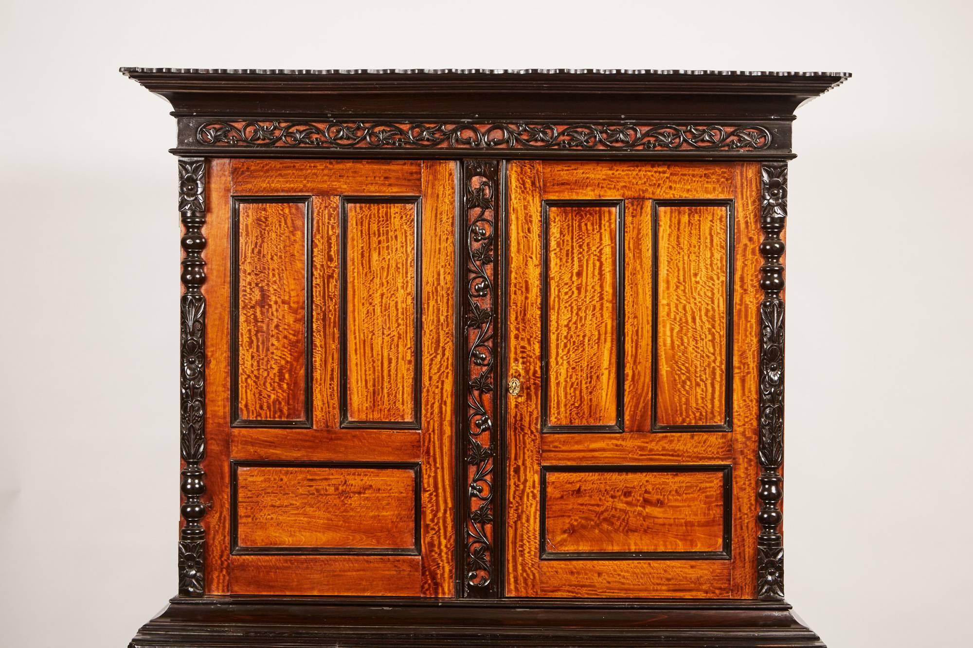 An exquisite 19th century British Colonial four-door cabinet in solid satinwood ebony and features remarkably beautiful carved molding designs of foliage throughout the exterior. The cabinet consists of an upper portion with two cupboard doors