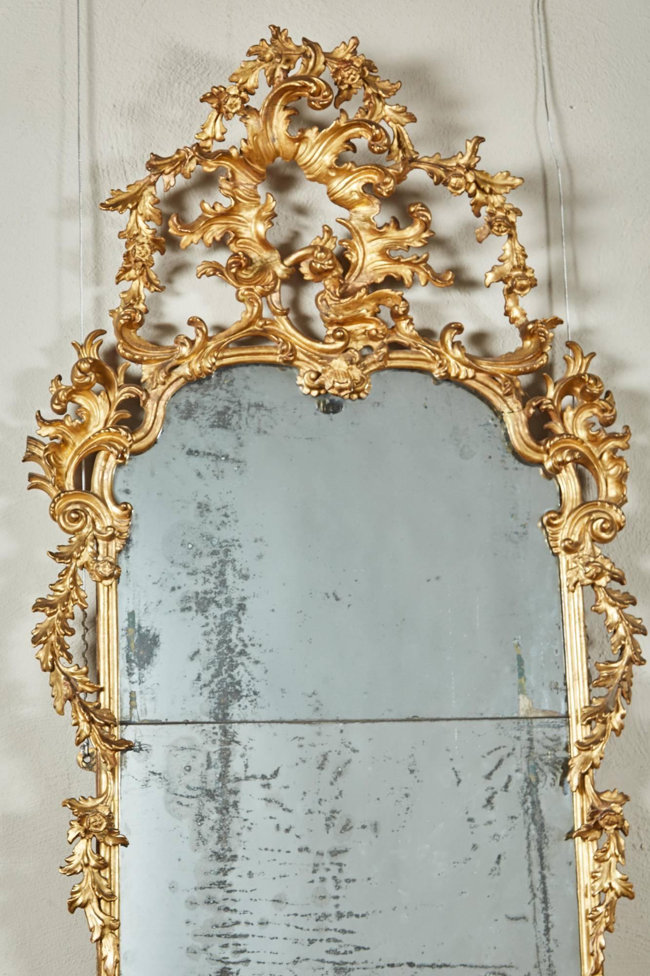 This giltwood Rococo mirror from mid-18th century Italy is a rectangular mirror surmounted by a large pierced rocaille with foliage garland extending downward toward simply decorated sides; the mirror plate in two sections; the lower frame section