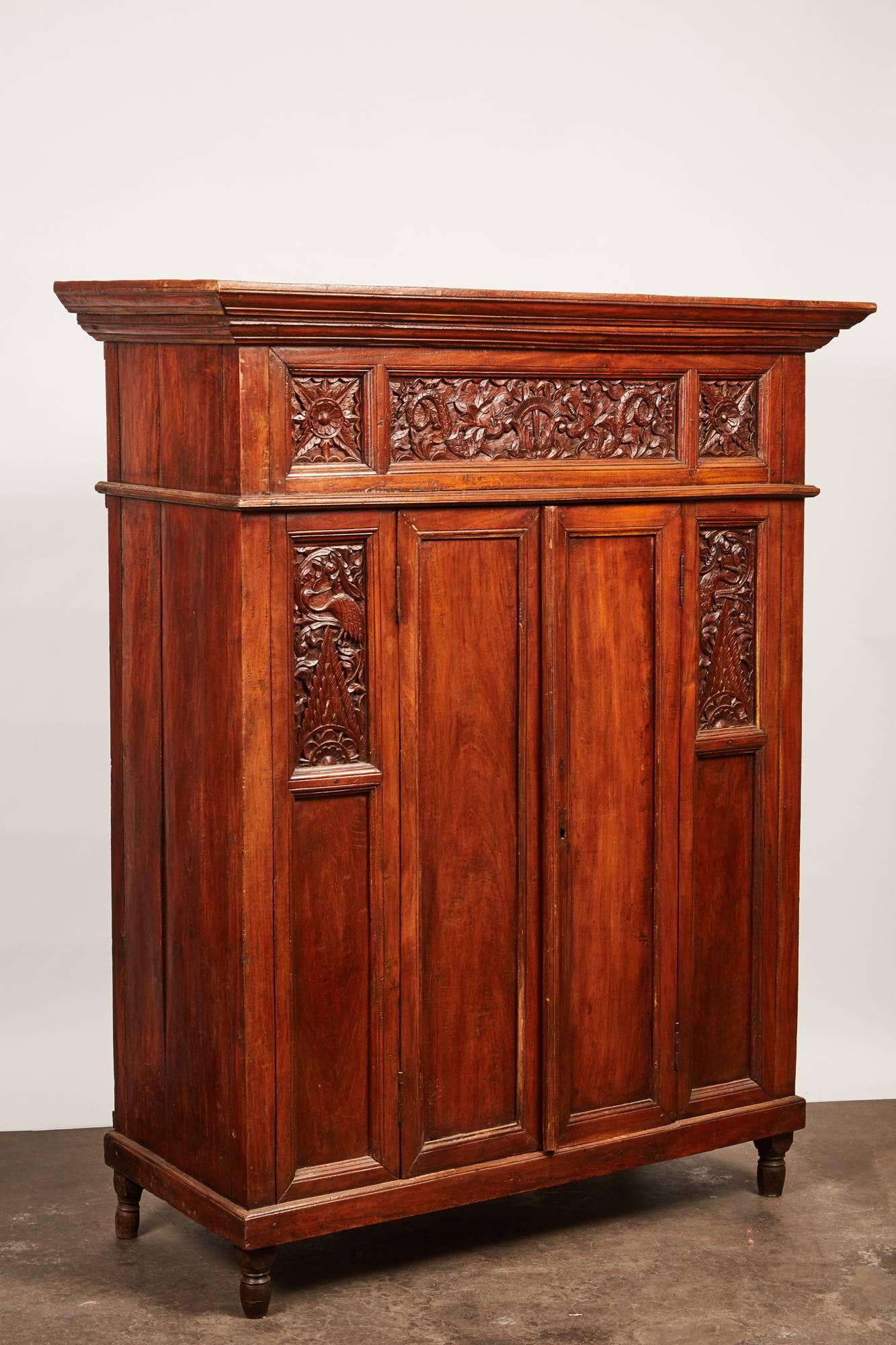 A unique Indonesian cabinet with a flaring cornice above a highly decorative frieze featuring complex carvings of foliage and dragon designs. Along the frame of the thin pair of doors there is another set of intricate carvings of peacocks and