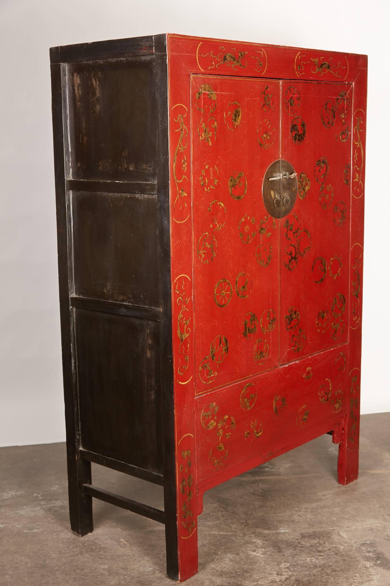 This 19th century Chinese Qing features a round face plate with pierced knobs and metal pulls. The cabinet is red lacquer with gold painted designs. The motifs on the facade are of dragons, lotus, butterflies, domestic life scenes, and cranes all in