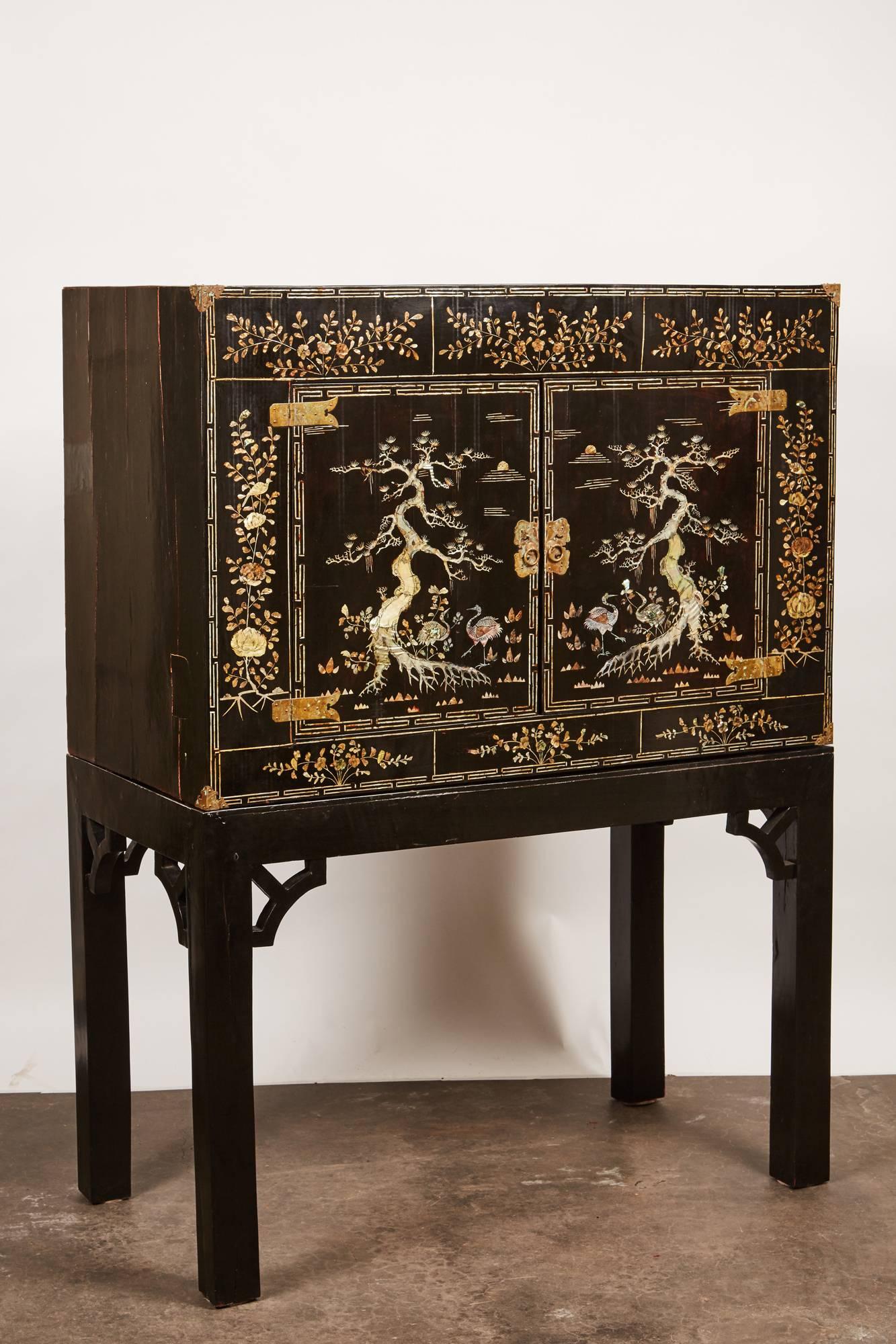 An early 20th century Vietnamese black two-door cabinet with mother-of-pearl detailing, resting on top of a black table stand. This Vietnamese piece is hinting at the Qing Dynasty by depicting motifs such as cranes, cypress trees, and peonies. The