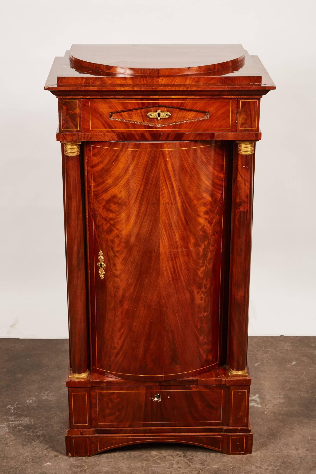 A remarkably elegant 19th century Empire cabinet that features beautiful crotch mahogany veneer. A drawer above one door that encloses three stationary shelves as well as a drawer below. A pair of columns flanks the curved door with gilt bases and