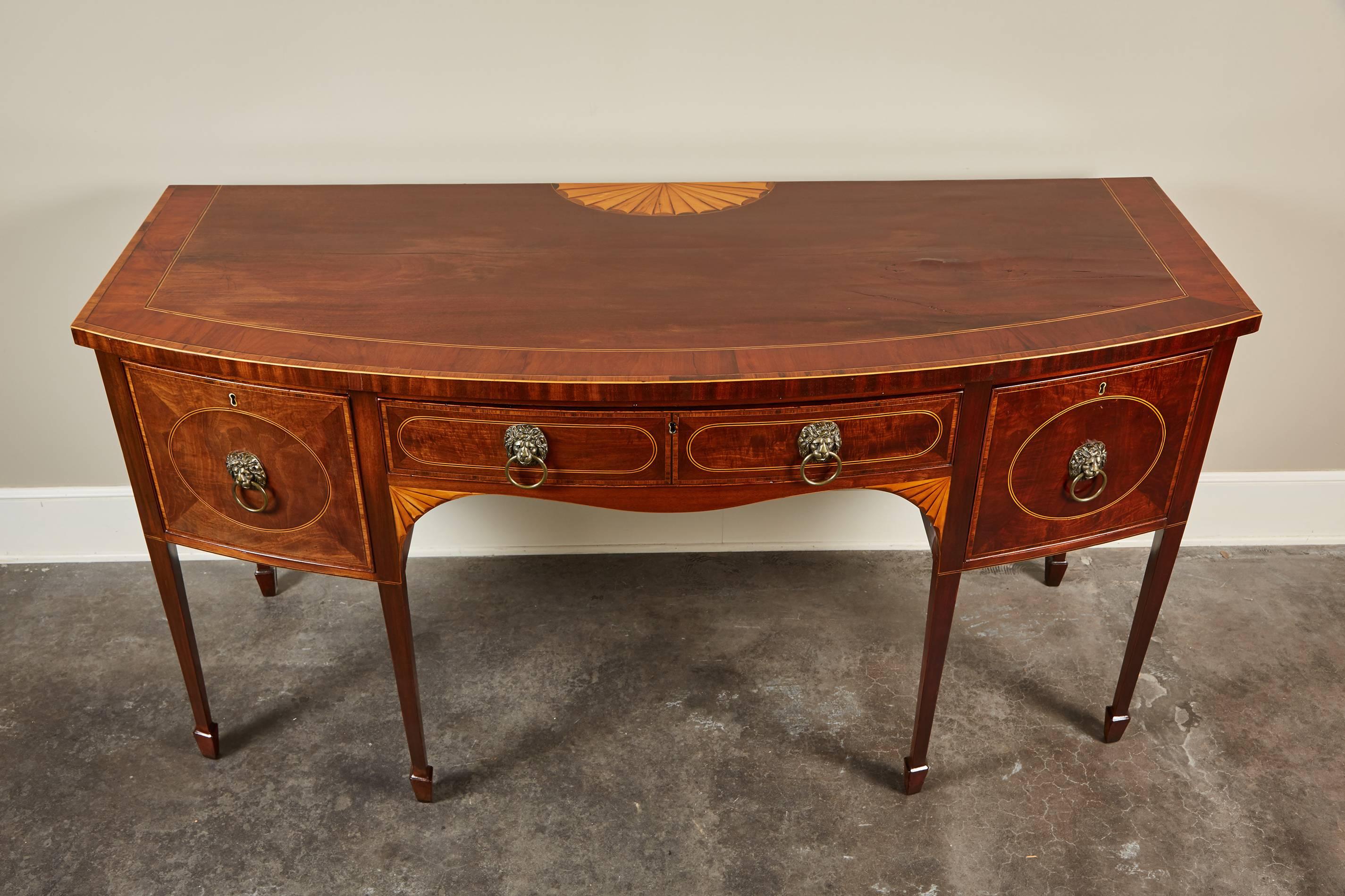 An early 19th century Georgian bow front sideboard in mahogany featuring inlaid fan detailing on the top and edge banding, sitting on spade feet. The sideboard also consists of two drawers and two doors with lion head hardware and oakwood interiors.