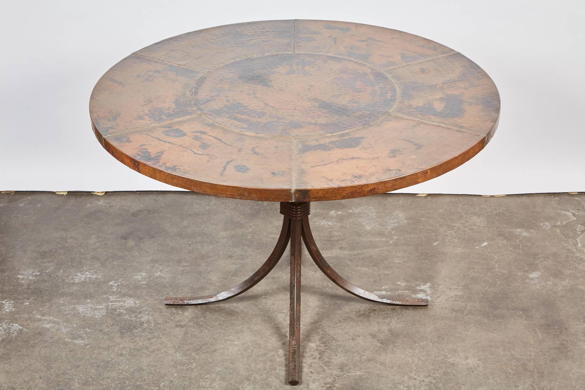 An Indian table that features a rust brown copper tabletop with seams sitting atop four C-Curved black iron legs bound together by a wrought iron twisted tie.