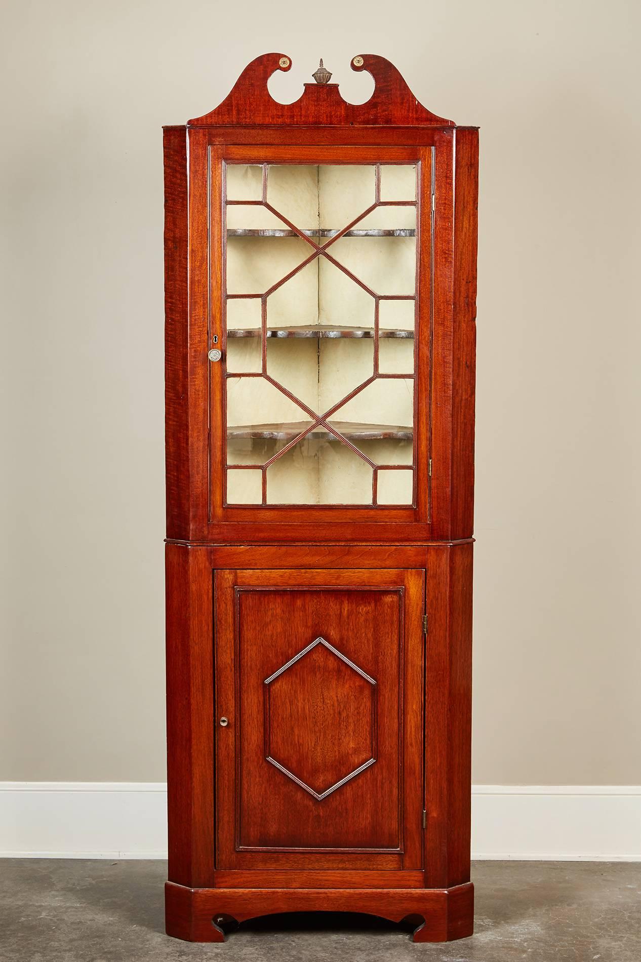 A 19th century Edwardian mahogany corner cabinet, with a swan pediment and a centered finial standing on bracket feet. The upper portion has an astragal glazed door enclosing three stationary shelving units, while the lower portion has an exterior