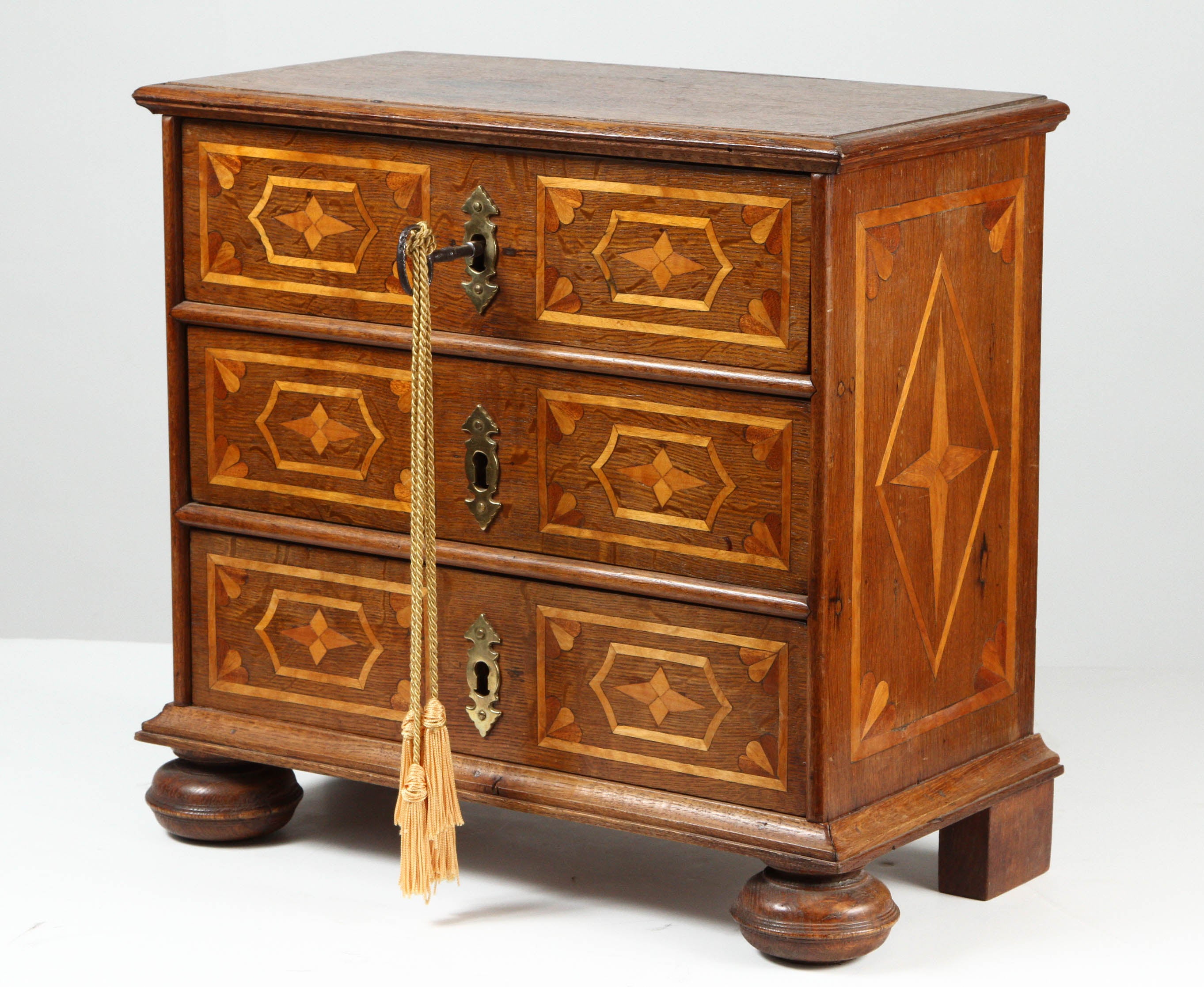 A miniature chest of drawers dating to the mid 19th century, once a sample carried by a traveling salesman.  The chest has three drawers, each of which has a keyhole.  Each drawer has inlaid decorations in a lighter shade on the left and right, and