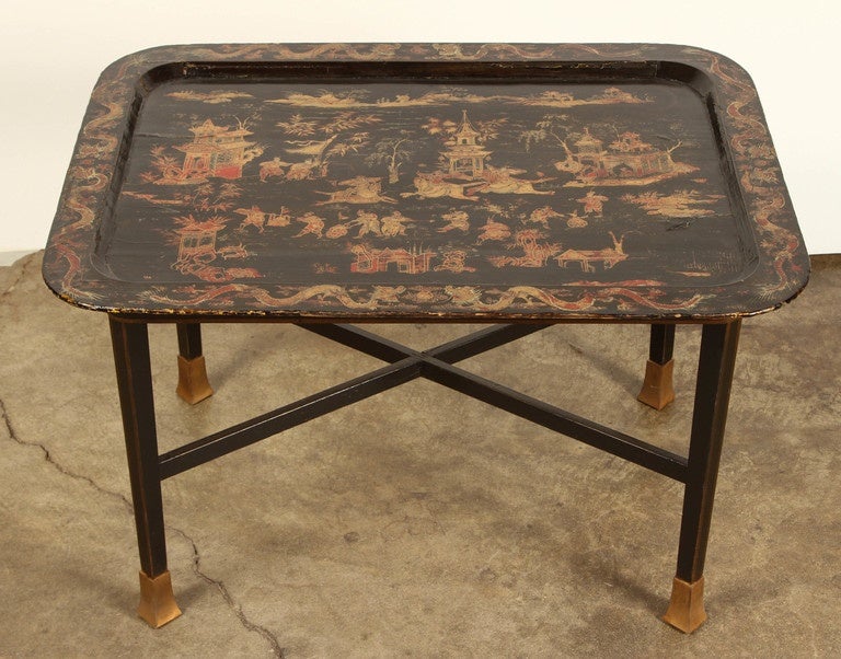 A 19th Century Chinoiserie tray table, painted with scenes of life including houses, horses, people and trees. The tray is mounted on four legs that are stabilized by an X stretcher. The legs terminate in brass sabot.