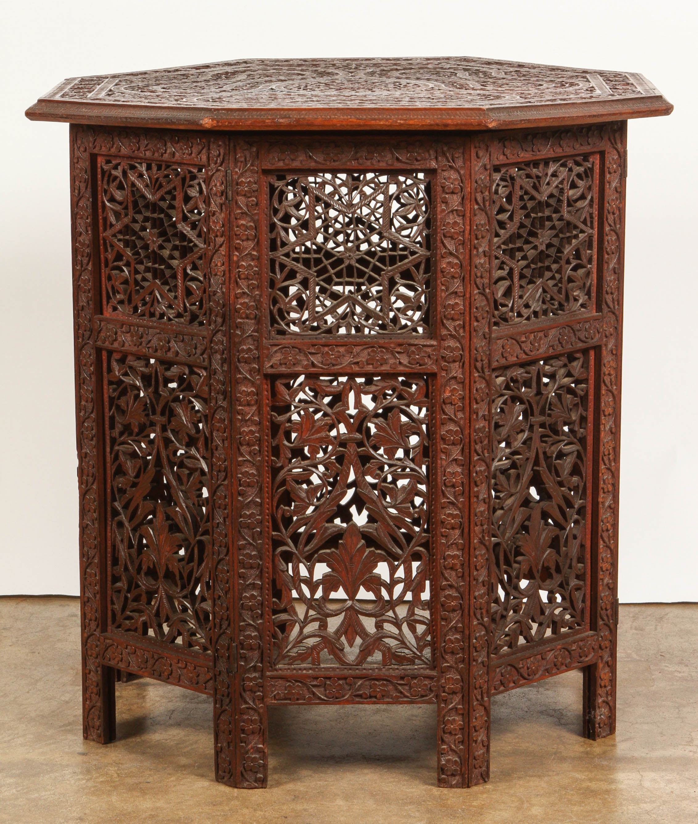 An eight-sided table from Syria in a red colored wood, elaborately and finely carved in leaf and floral patterns, as well as a row of hexagon shapes at the top of the base.