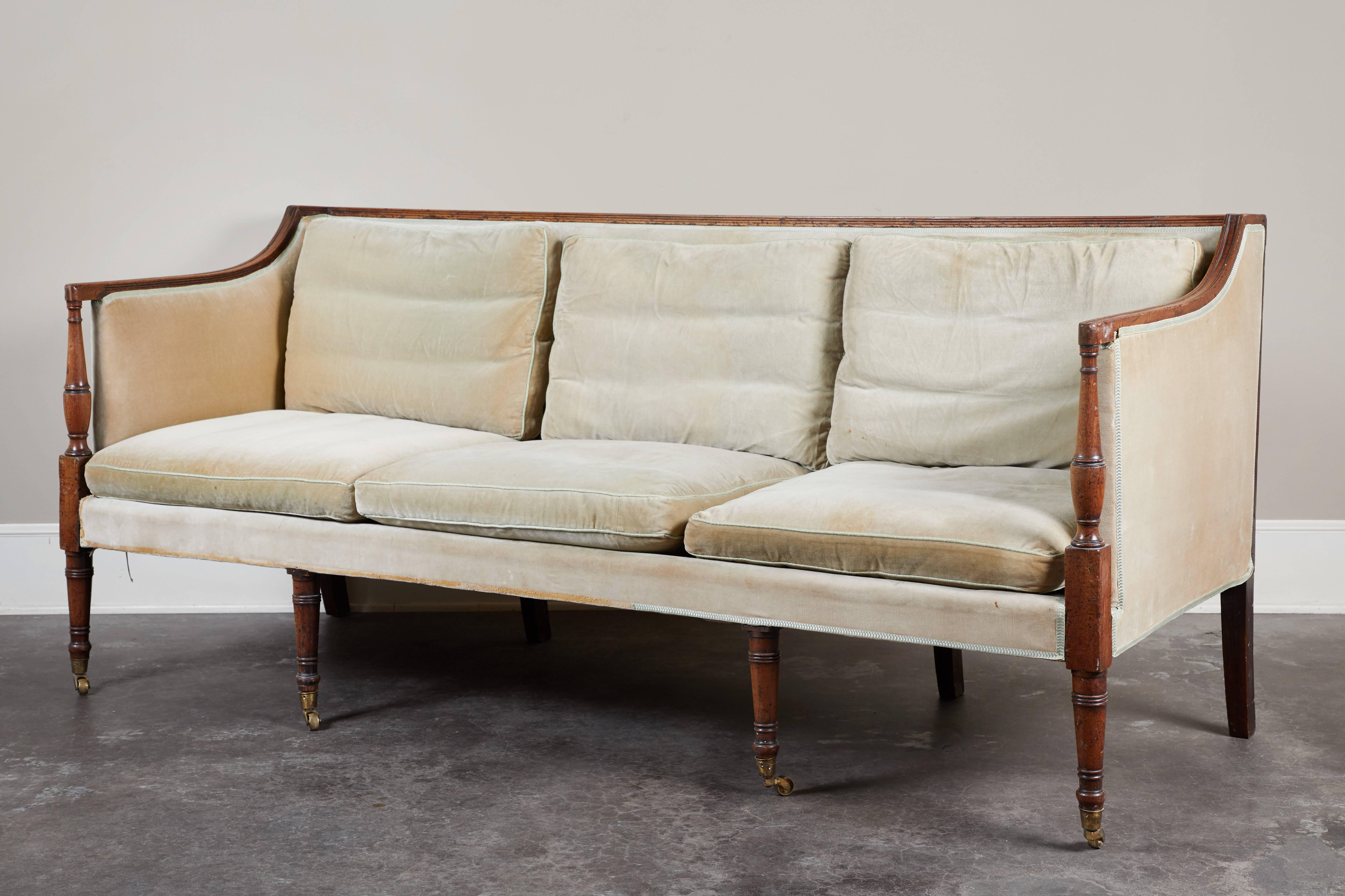 An early 19th century English Regency mahogany-trimmed sofa.  Turned front legs on wheels, delicate armrests and transitional, square shape.