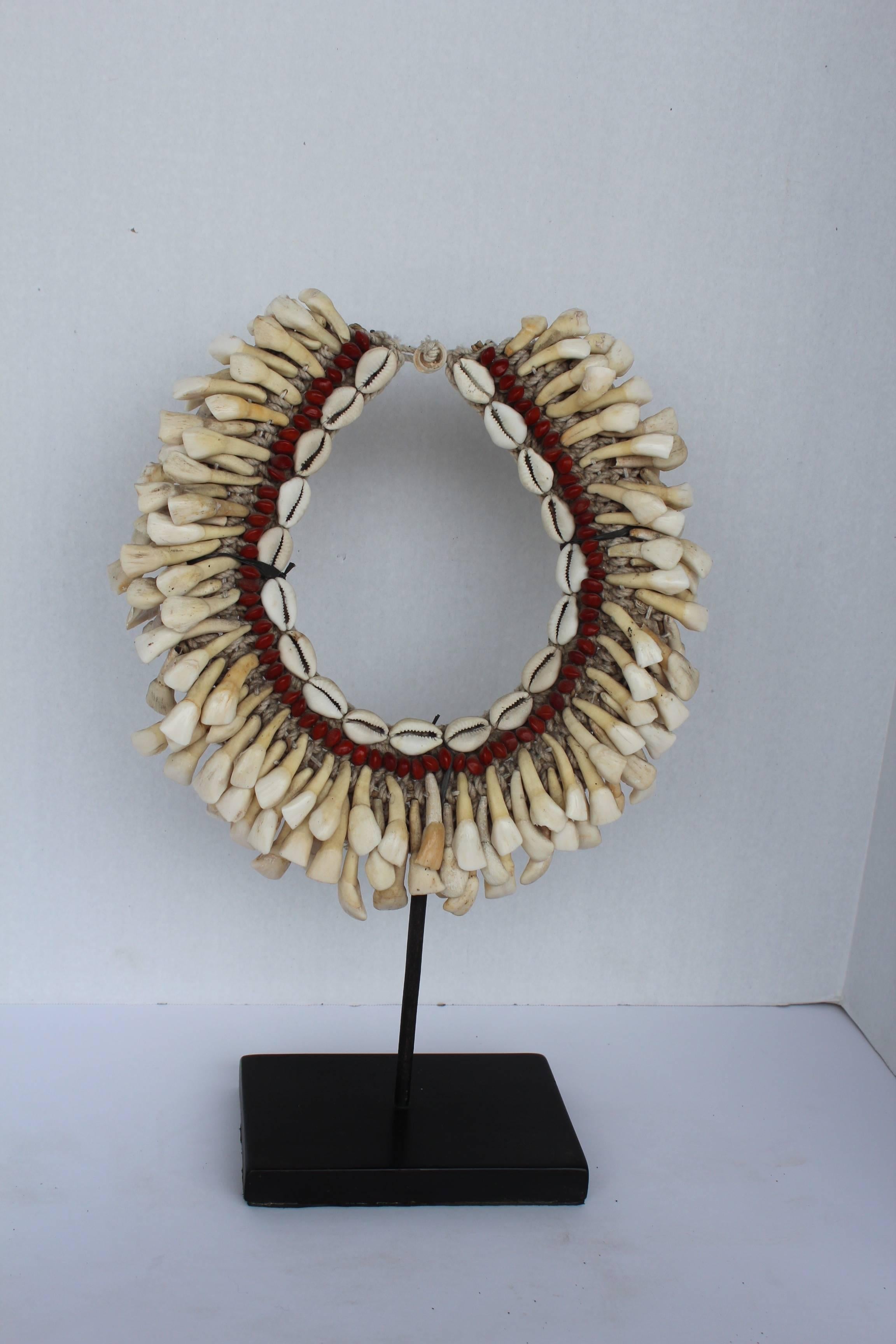 Three layers of antique buffalo teeth hand tied on handwoven hemp fiber collar with central band of red wood beads and cowry shells as necklace attached to custom stand. 

Buffalo teeth was once used as a form of currency or the bribe would wear it