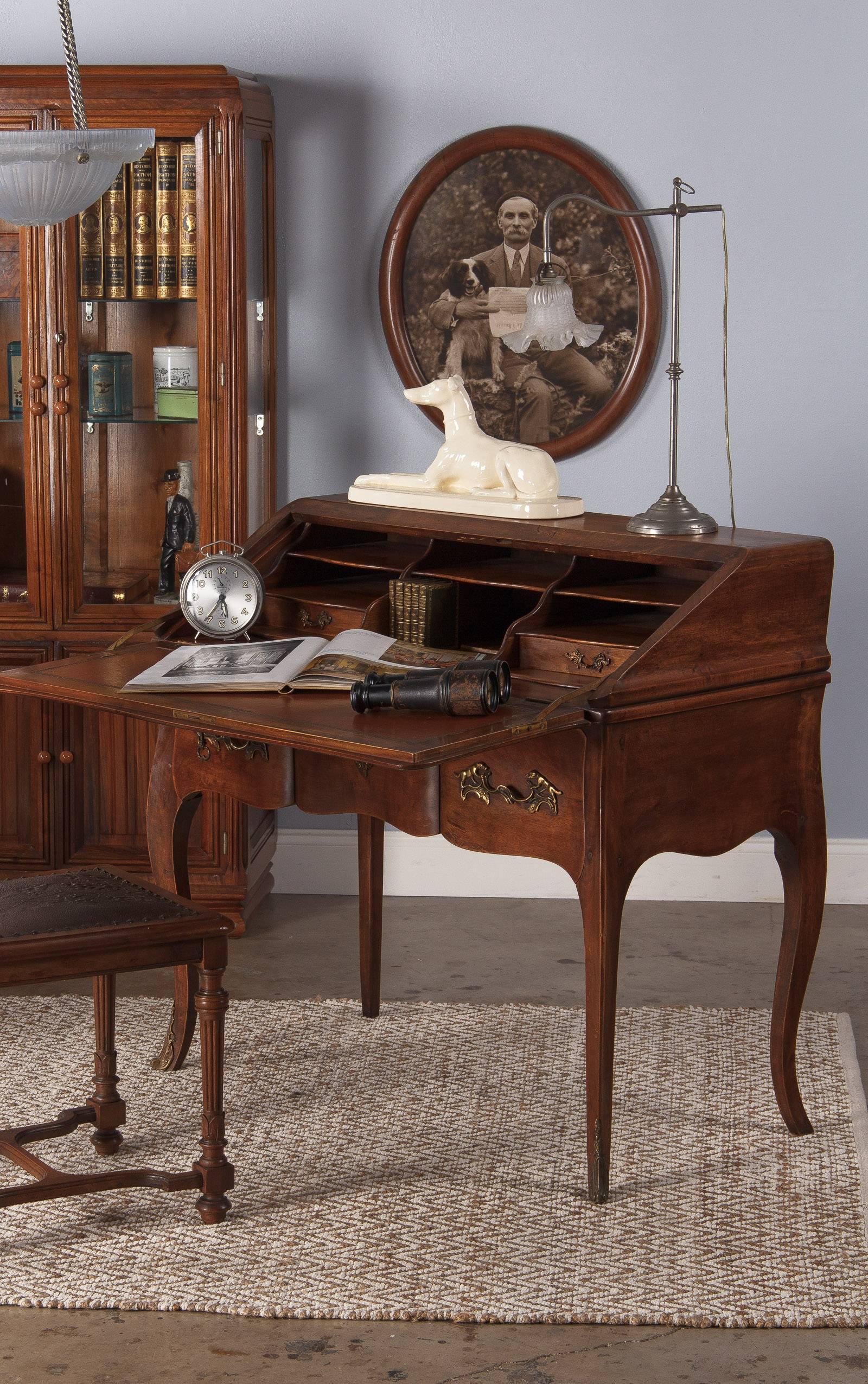 A handsome Secretaire desk made of walnut in the Louis XV style, circa 1920s. A brass key opens the drop front to an embossed tobacco leather top and reveals six storage compartments and two small drawers in golden oak. The back center features a