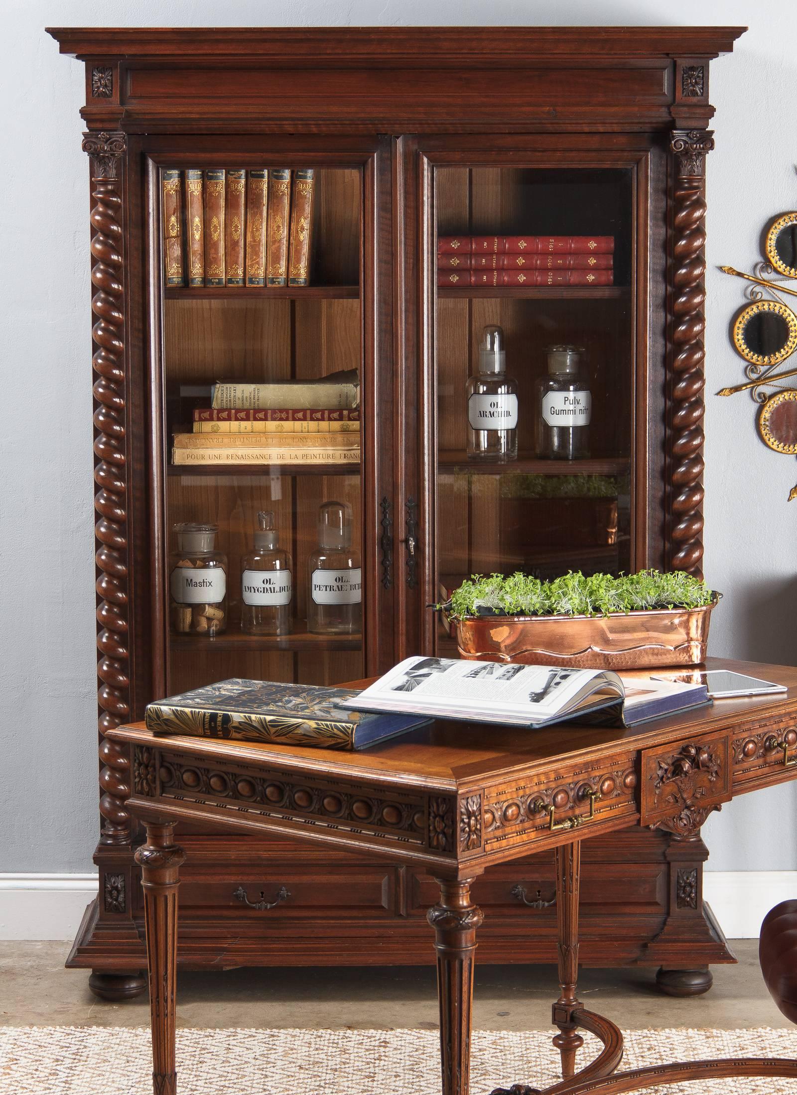 A comely walnut bookcase in the Louis XIII style, circa 1880. Either side is graced by an elegant barley twist with a Corinthian capital at the top. Two drawers with black iron drop handles sit over a heavily carved molded spreading plinth. The