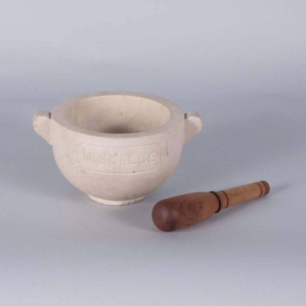 A French Apothecary marble mortar with a wooden pestle purchased in the Provence Region. The marble has a carved label with the inscription M. Sollce on one side and C.D on the other. Beige in color with thin, sparse veins running throughout the